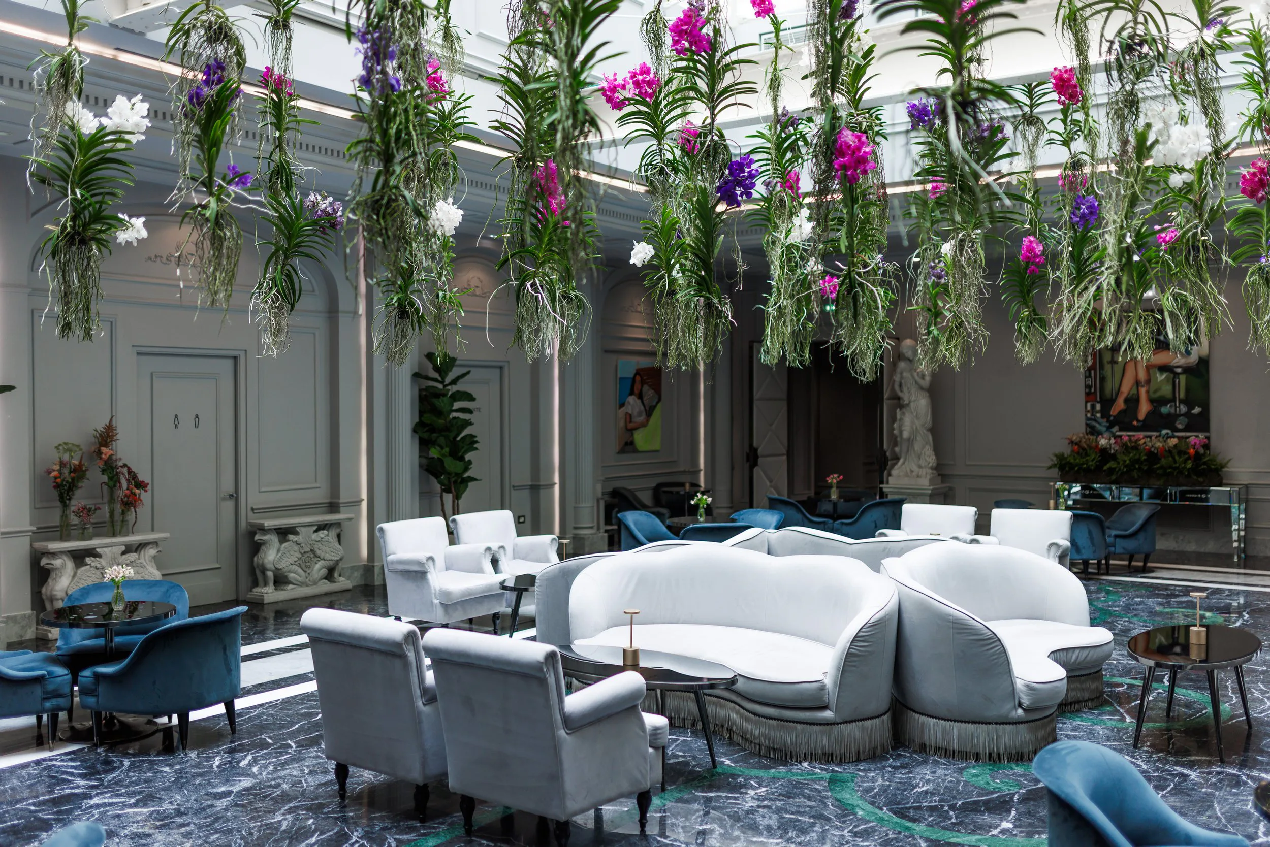 Salon élégant avec mobilier blanc et bleu, plantes en pot, sculptures et éclairage naturel, avec des fleurs suspendues au plafond.