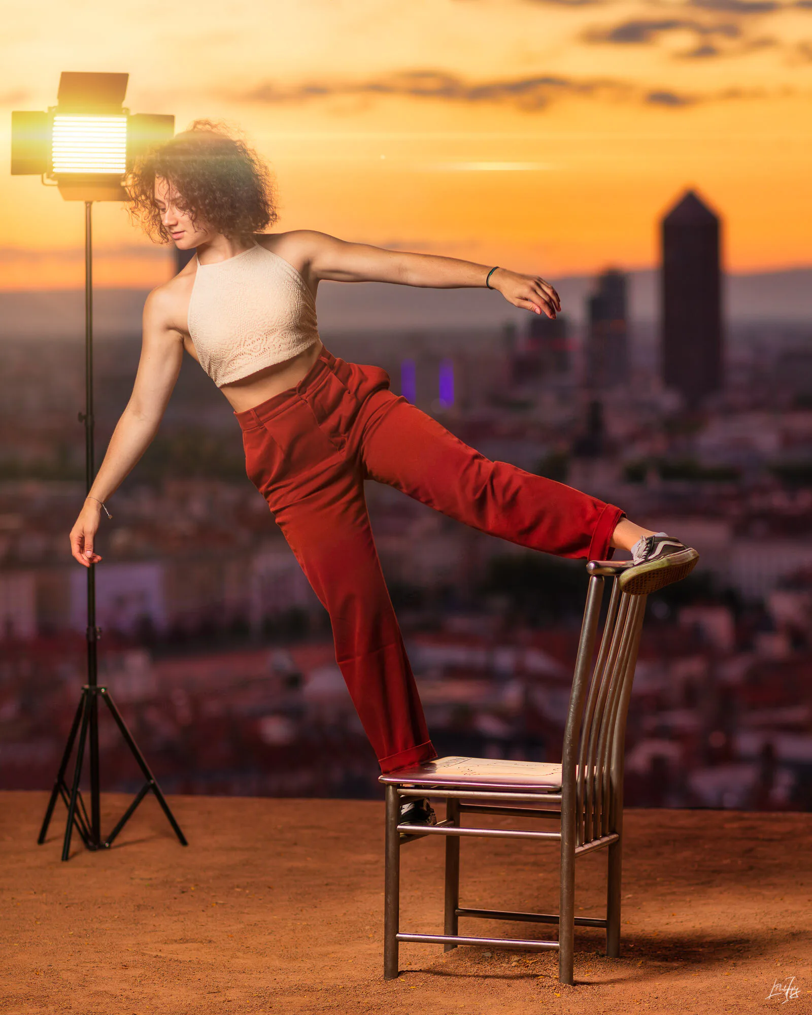 Une femme debout sur une chaise, en équilibre avec un pied, dans un studio avec un fond extérieur de coucher de soleil sur une ville.