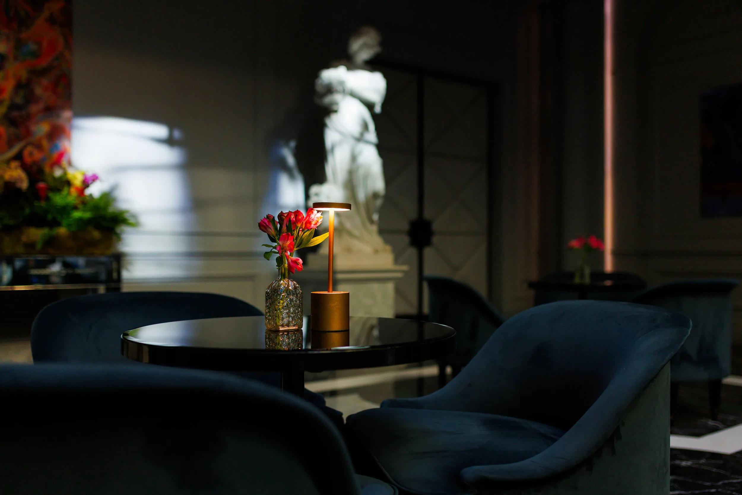 Intérieur d'une salle élégante avec un coin salon, un vase avec des fleurs roses, et une petite lampe sur la table. En arrière-plan, une sculpture blanche et des murs ornés.