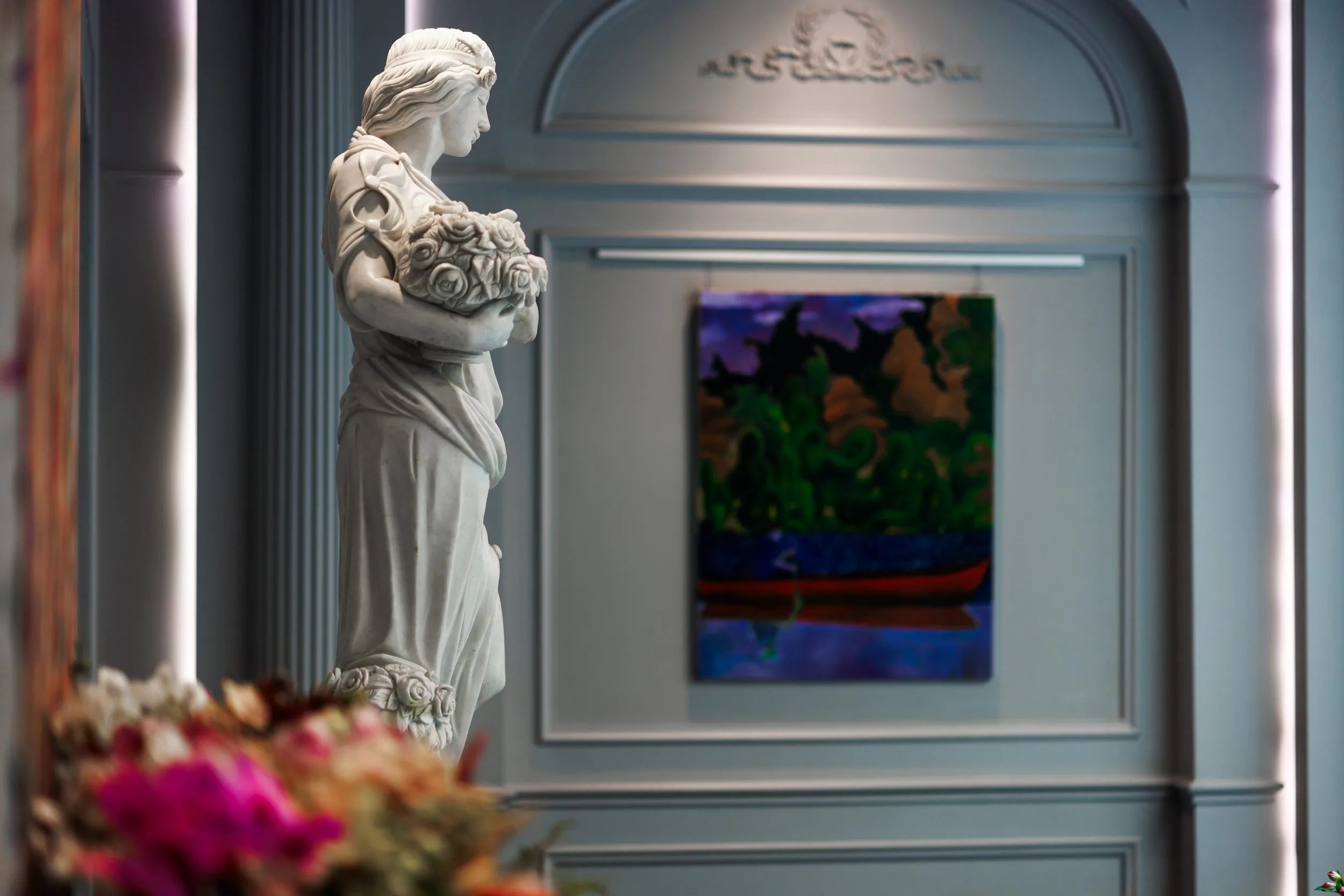 Une sculpture en marbre représentant une femme tenant un bouquet de roses, située dans une salle d'exposition avec un tableau coloré en arrière-plan.