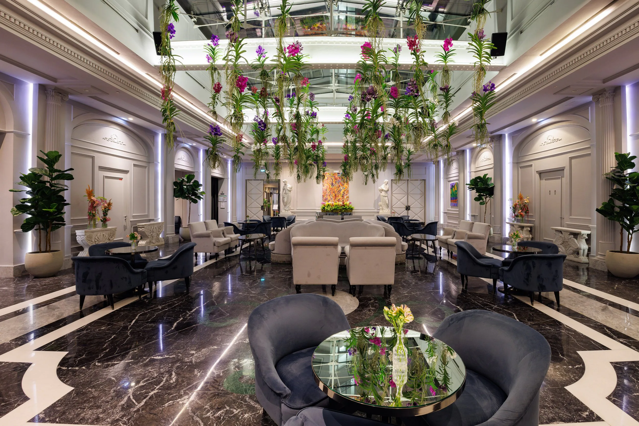 Un hall élégant avec un plafond en verre et des plantes suspendues, des statues blanches, des fauteuils et des tables, décor floral, intérieur lumineux avec éclairage moderne.