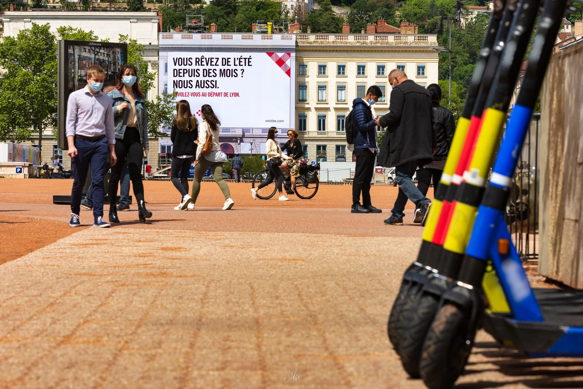 Trottinette et passants place Bellecour, bâche Volotea en arrière-plan