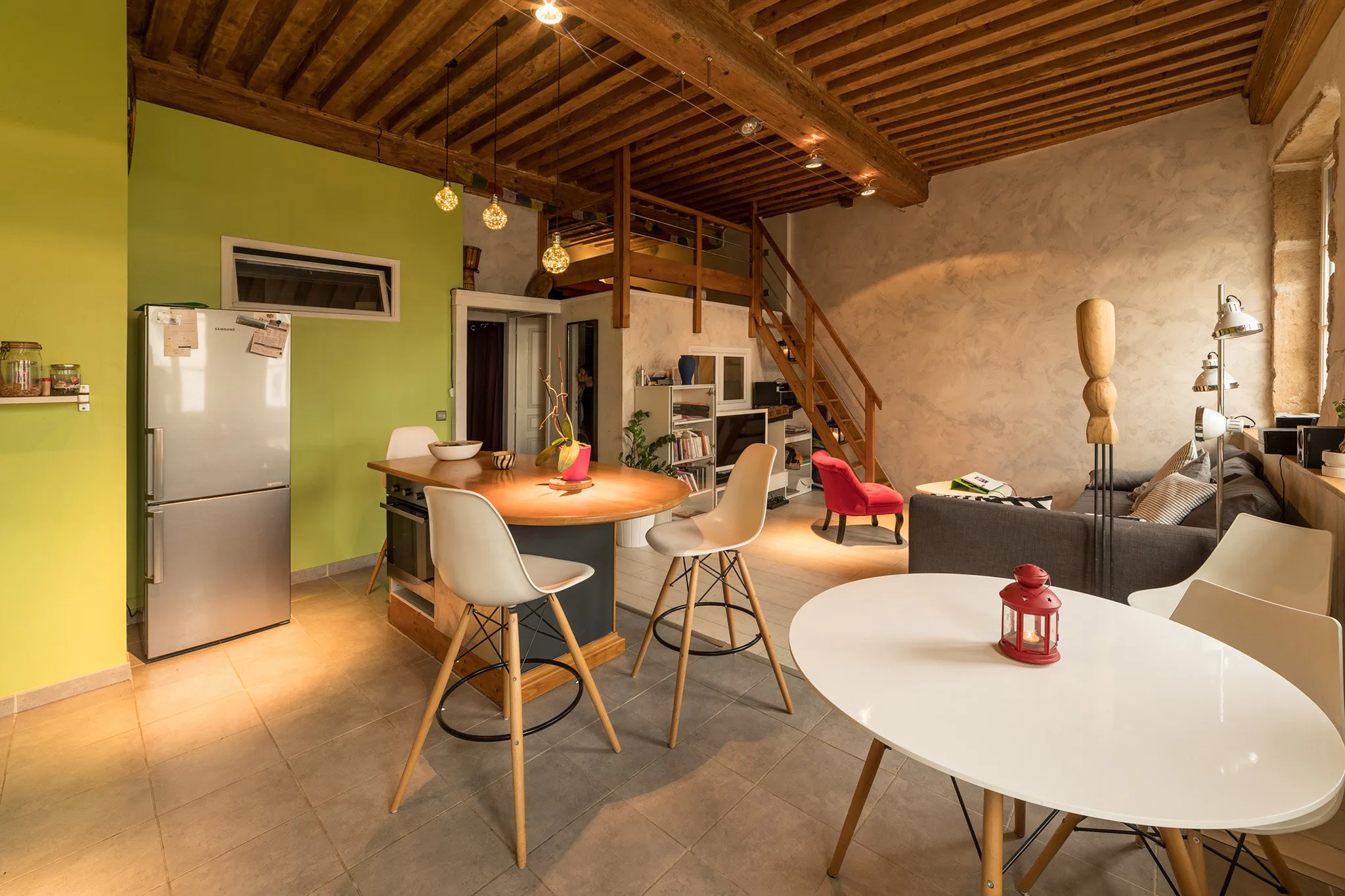 Intérieur d'un salon et cuisine avec murs verts et beige, plafond en bois, mobilier moderne, lampes suspendues, escaliers en bois, canapé gris, fauteuil rouge, tables blanches, décorations variées.
