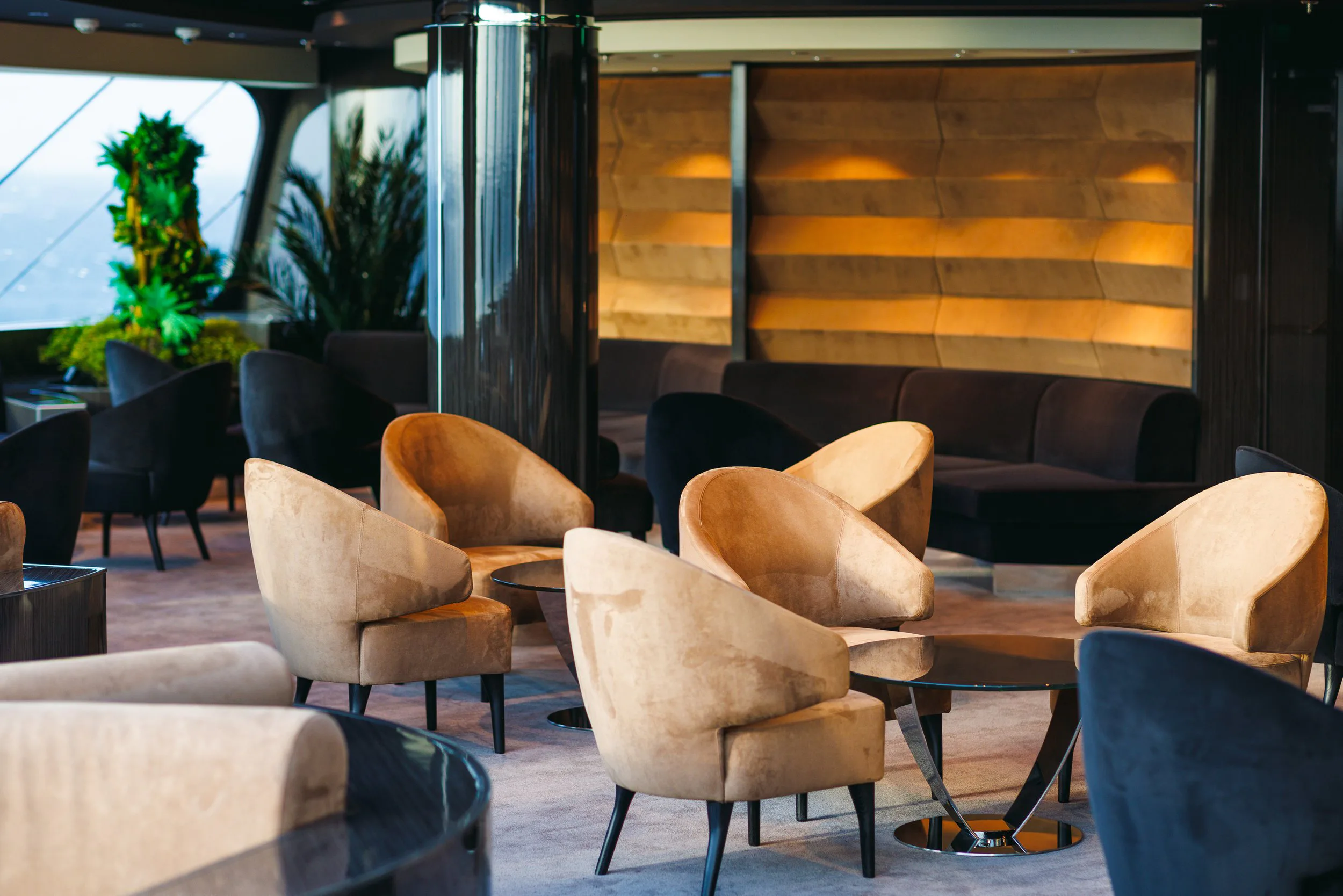 Salon de lounge élégant avec fauteuils en velours beige et sciés en bois foncé autour de petites tables en verre, lumière tamisée, plantes vertes et fenêtres donnant sur l'extérieur.