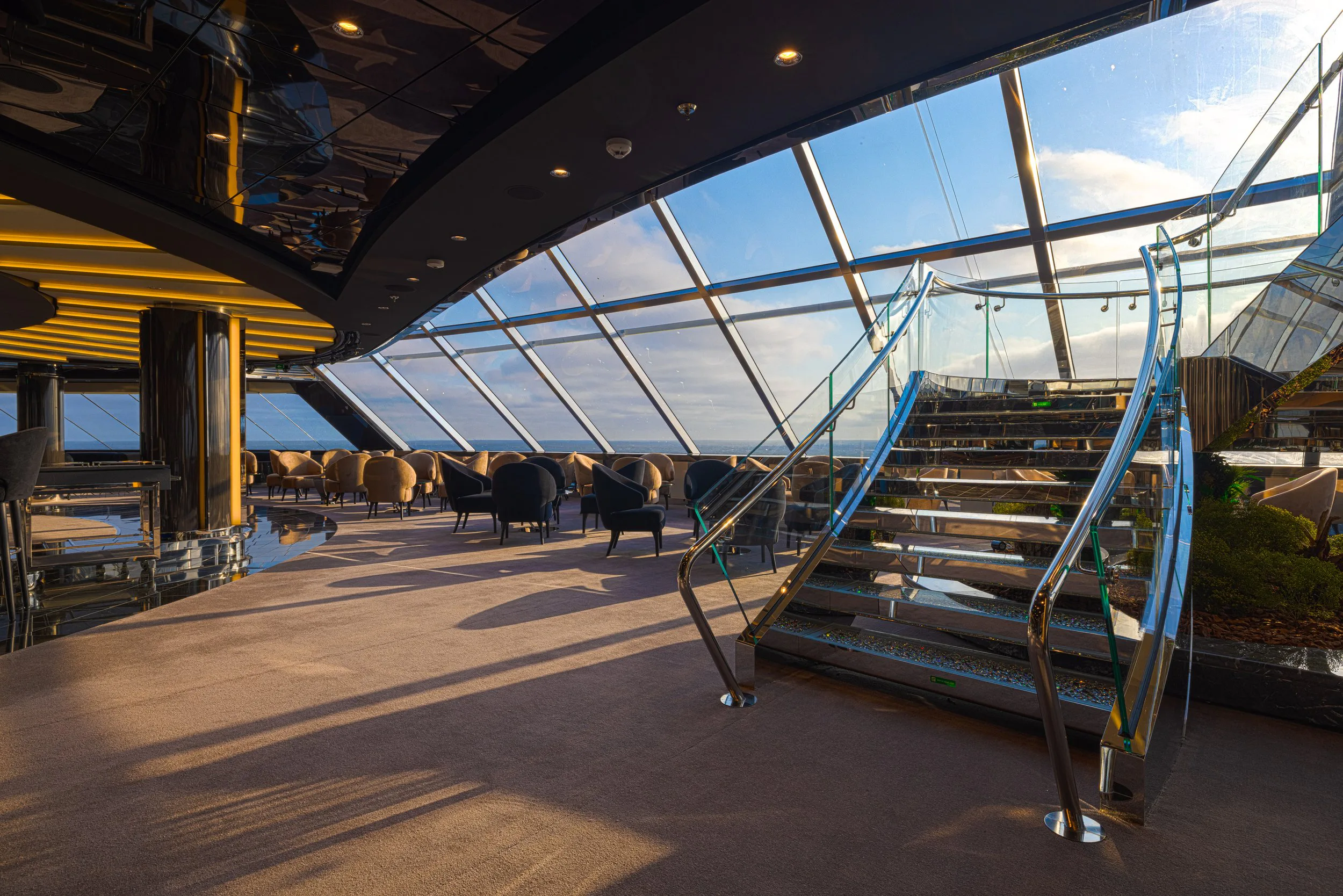 Intérieur d'un espace lounge avec des sièges modernes, des escaliers en verre, et de grandes fenêtres offrant une vue sur le ciel et l'océan.