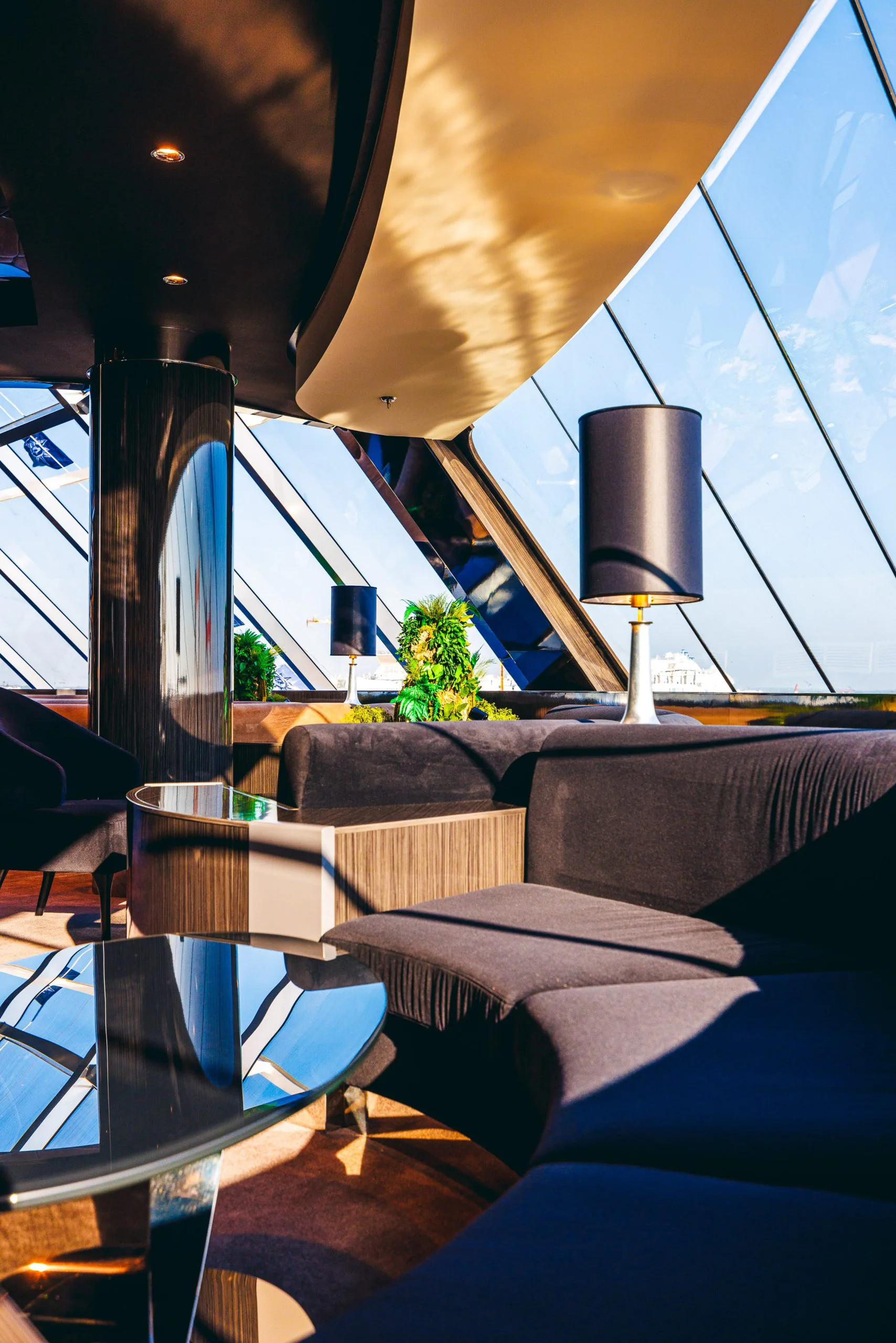 Intérieur moderne avec canapé noir, lampes noires sur tables, plantes vertes, grandes fenêtres en verre avec un ciel bleu visible, et un mobilier élégant.