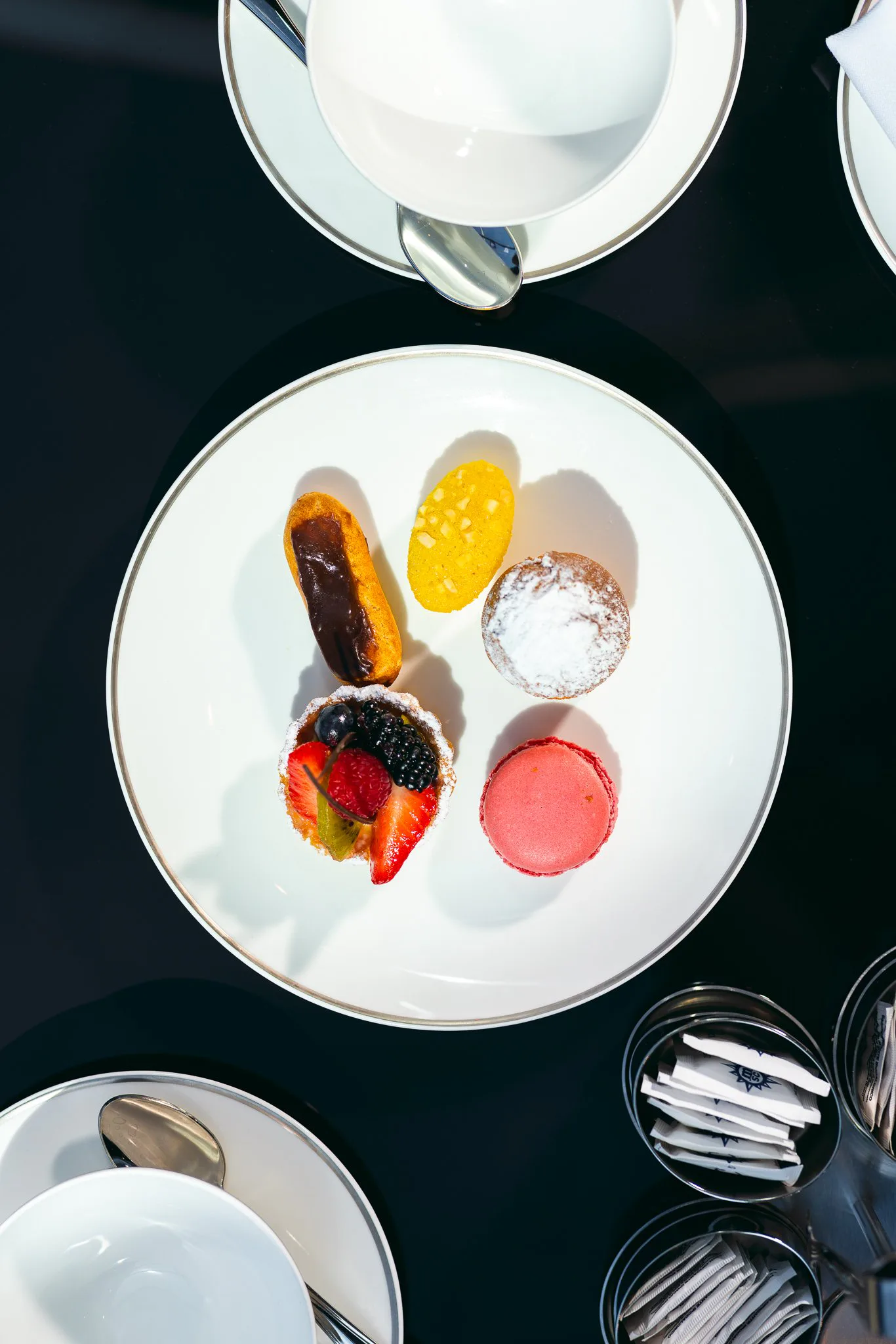 Un plateau avec plusieurs petits desserts, dont une fraise couverte de chocolat, une tarte aux fruits, un macaron rose, un biscuit en poudre, un biscuit jaune, un biscuit éclat de sucre, et un petit gâteau en pâte à sucre à décor fruitier.