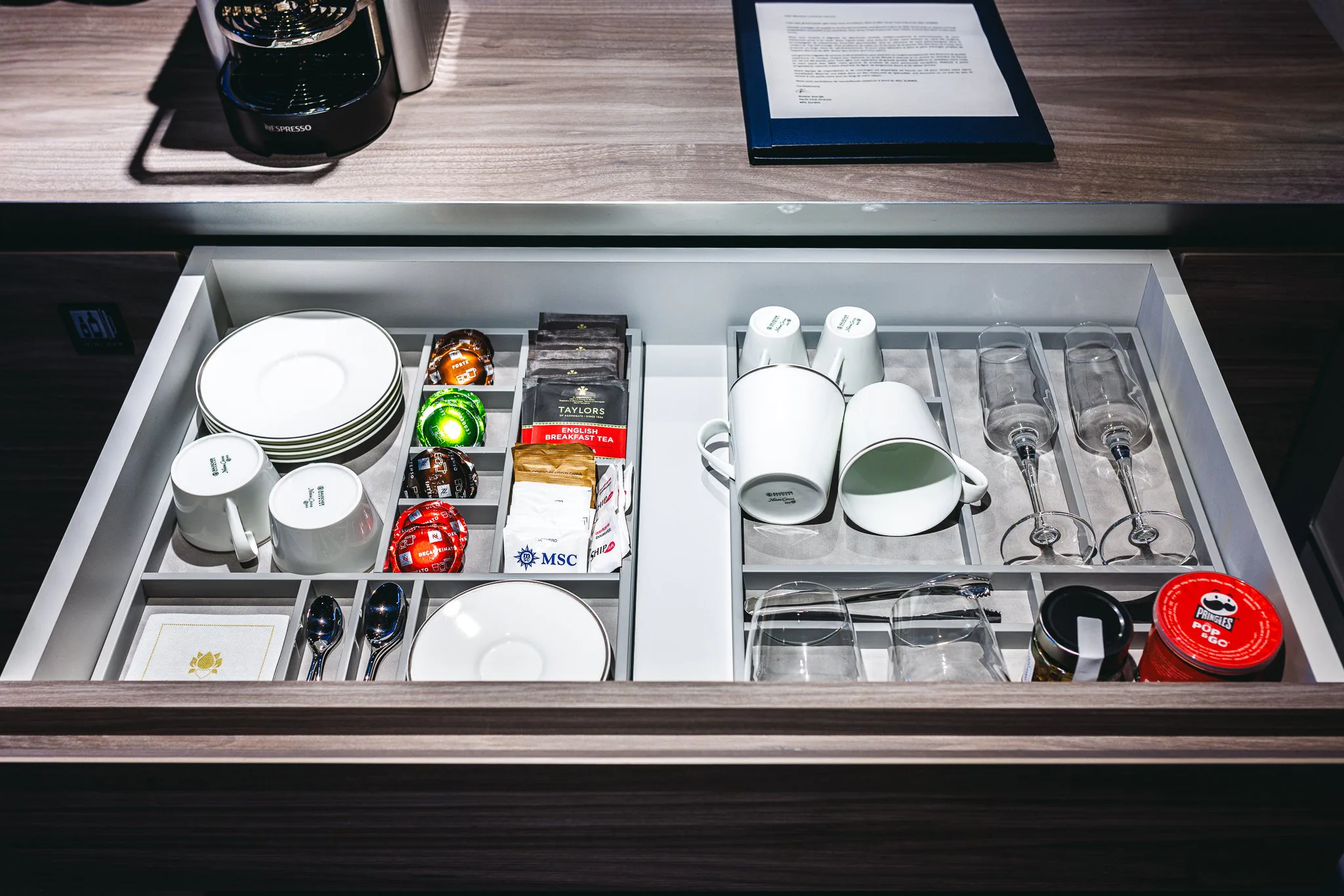 Coffre de chambre d'hôtel contenant des tasses, des assiettes, des sachets de thé, des capsules de café, des verres à vin, et des aliments comme des gelées et une confiture.