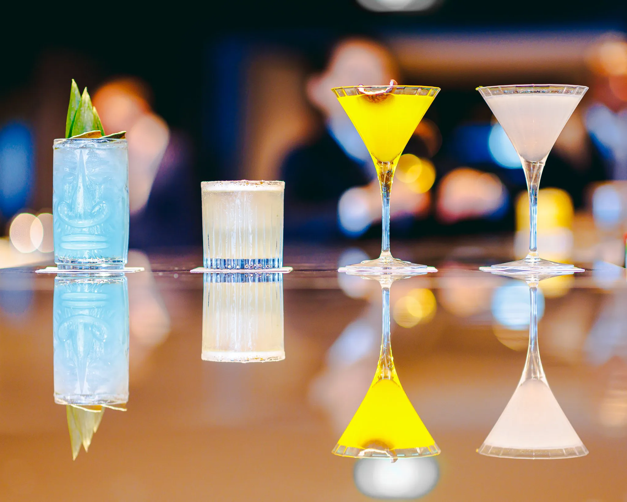 Cinq cocktails colorés alignés avec leurs reflets sur une surface en bois dans un bar, fond flou avec des lumières chaudes.