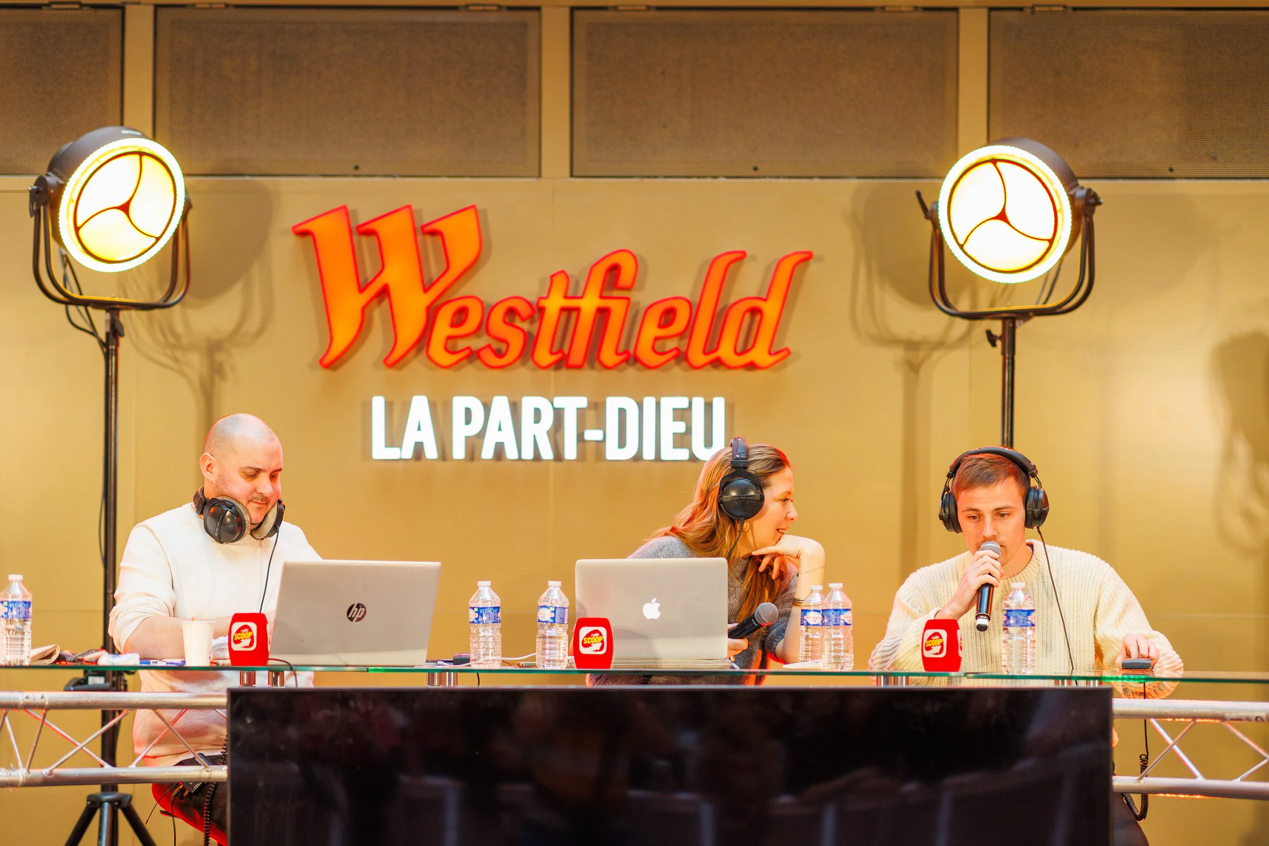 Trois personnes assises à une table discutant lors d'une émission ou d'une réunion. Deux hommes et une femme, portant des casques audio, avec des microphones et des bouteilles d'eau. En arrière-plan, un panneau avec le texte 'Westheld La Part-Dieu'.