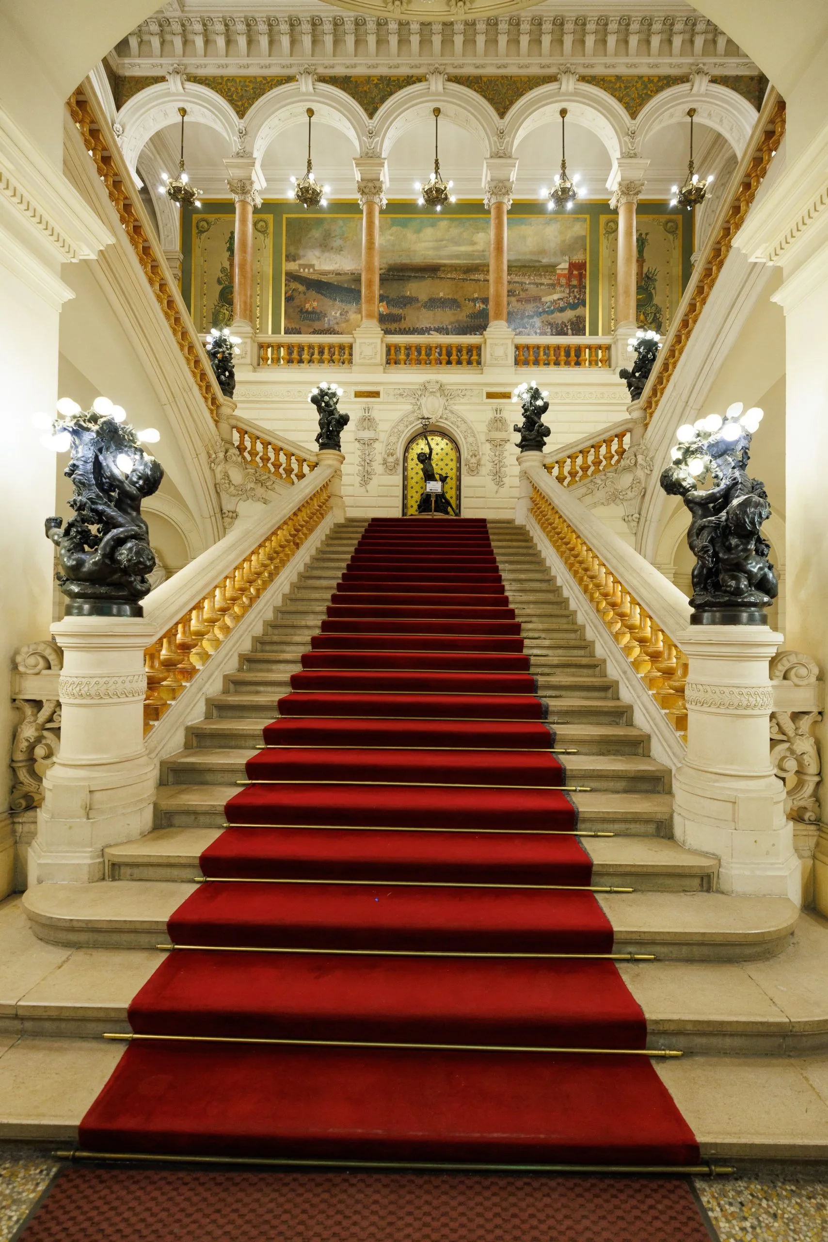 Escalier royal avec tapis rouge, statues en bronze lumineux, et plafond orné dans un hall d'hôtel ou palais élégant.