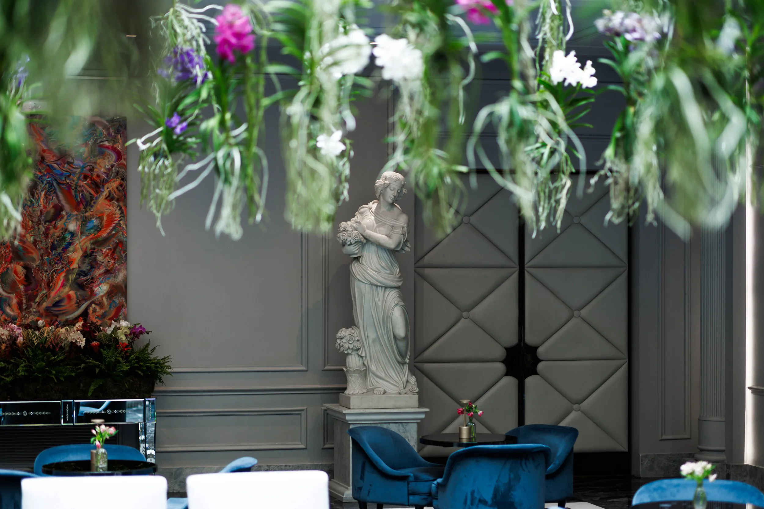 Intérieur élégant avec une statue antique de femme en pierre, entourée de mobilier de salon bleu, plusieurs petites tables avec des vases de fleurs, murs gris avec moulures, décorations d'art moderne et plantes suspendues avec fleurs