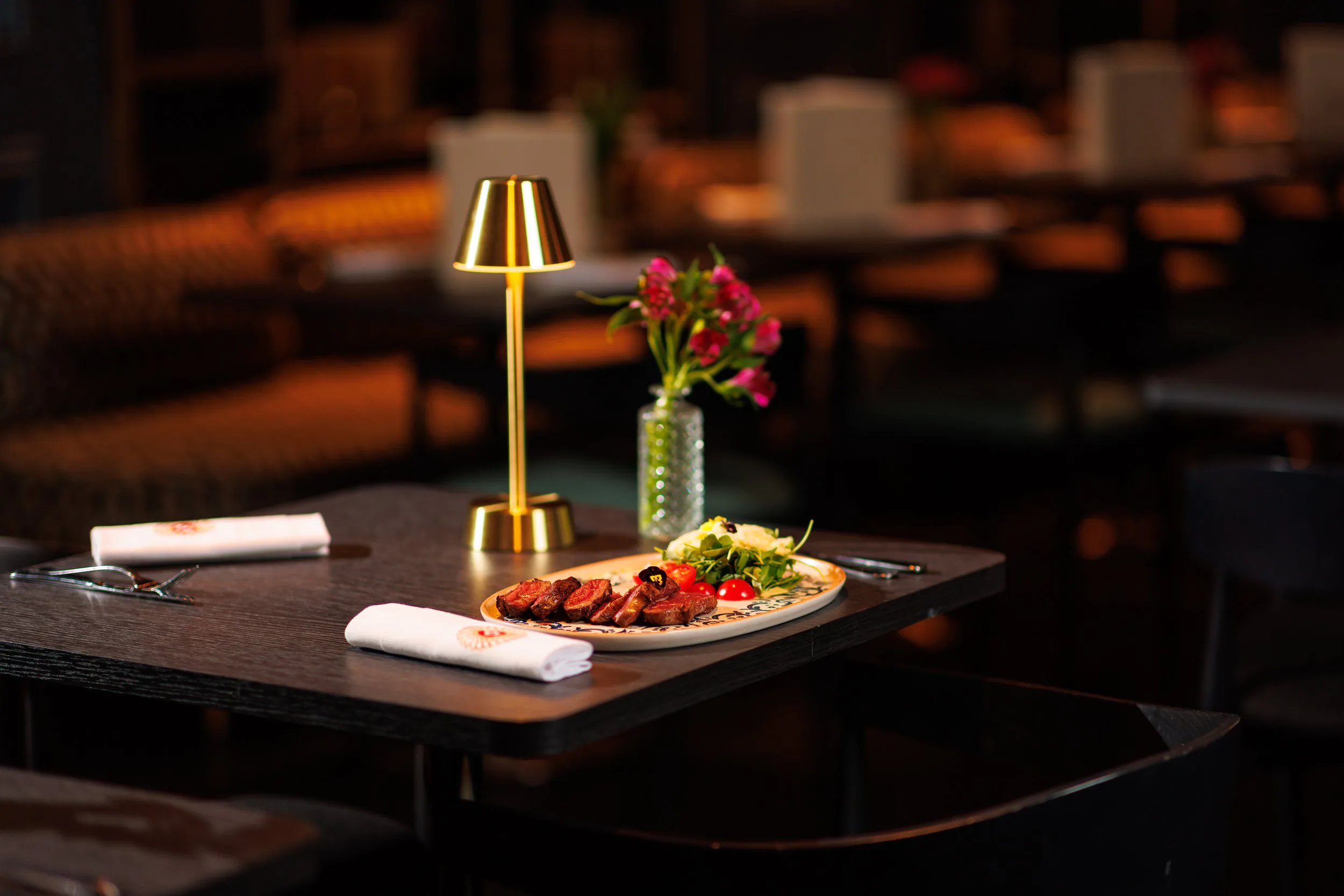 Une table d'un restaurant élégamment dressée avec un plat de viande, une salade et des tomates cerises, une lampe en or et un vase avec des fleurs roses, dans un cadre sombre et sophistiqué.