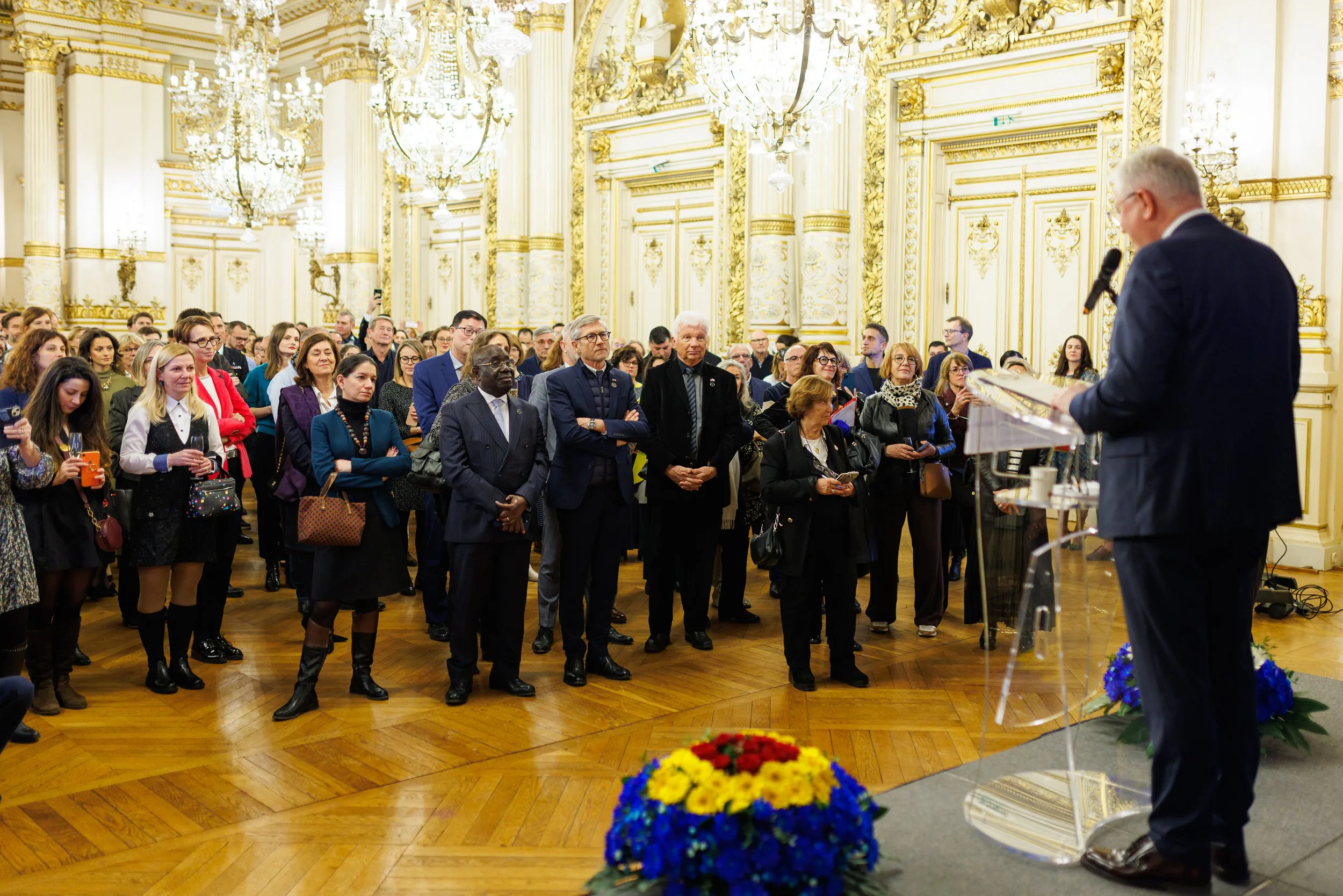 Une conférence ou un discours dans une salle ornée de dorures et de lustres en cristal, avec un homme en costume parlant au micro face à un public diversifié, composé d'hommes et de femmes attentifs, certains prenant des photos ou vidéos.