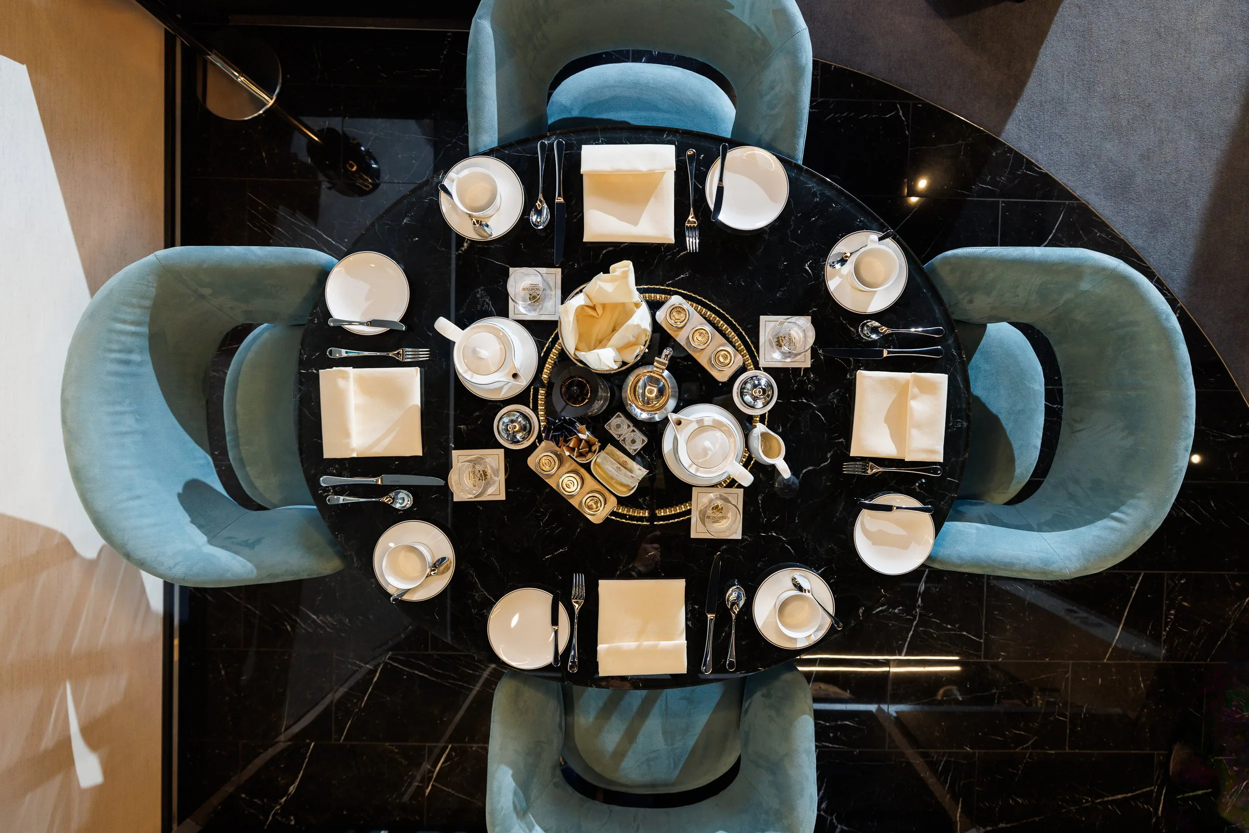Vue du dessus d'une table de petit-déjeuner avec assiettes, tasses, couverts, et serviettes, entourée de six chaises en velours bleu clair.