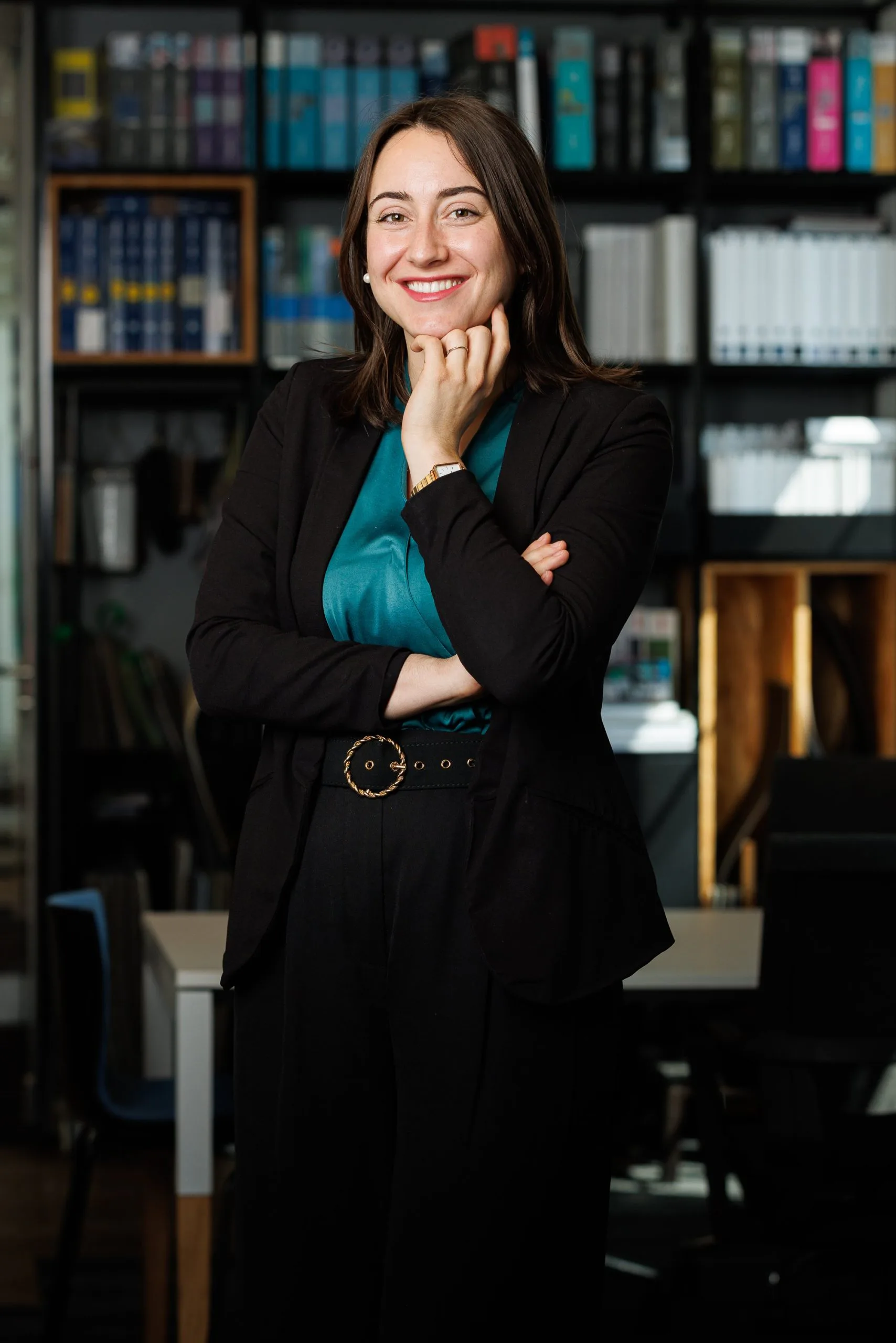 Femme souriante en tenue professionnelle pose dans un bureau avec des étagères remplies de livres en arrière-plan.