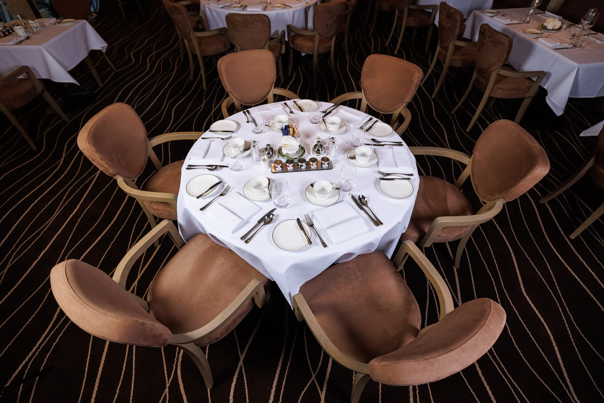 Table ronde avec nappe blanche, décorée pour un repas, entourée de chaises en velours brun, dans une salle de banquet.