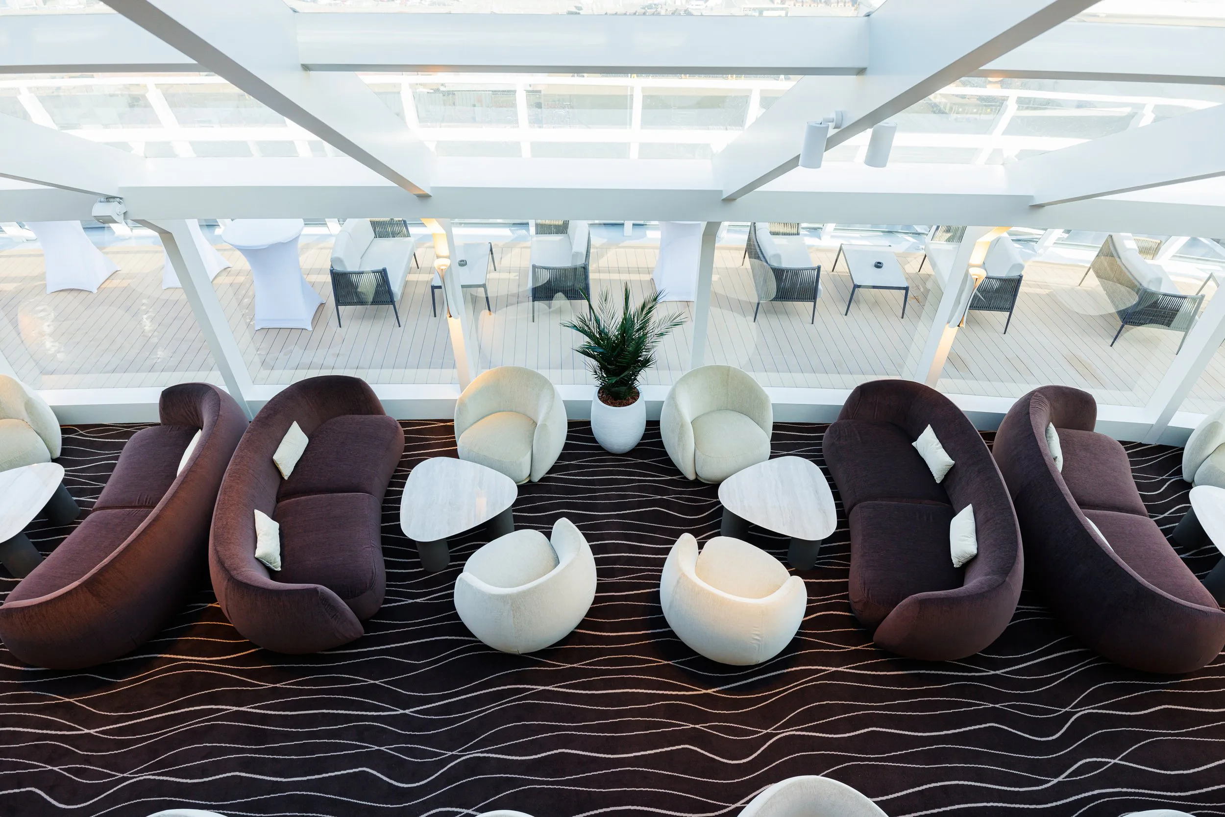 Salle de lounge moderne avec des canapés beiges et marron, des fauteuils arrondis, tables basses, et un plafond en verre laissant entrer beaucoup de lumière naturelle, avec un espace extérieur visible par la fenêtre.