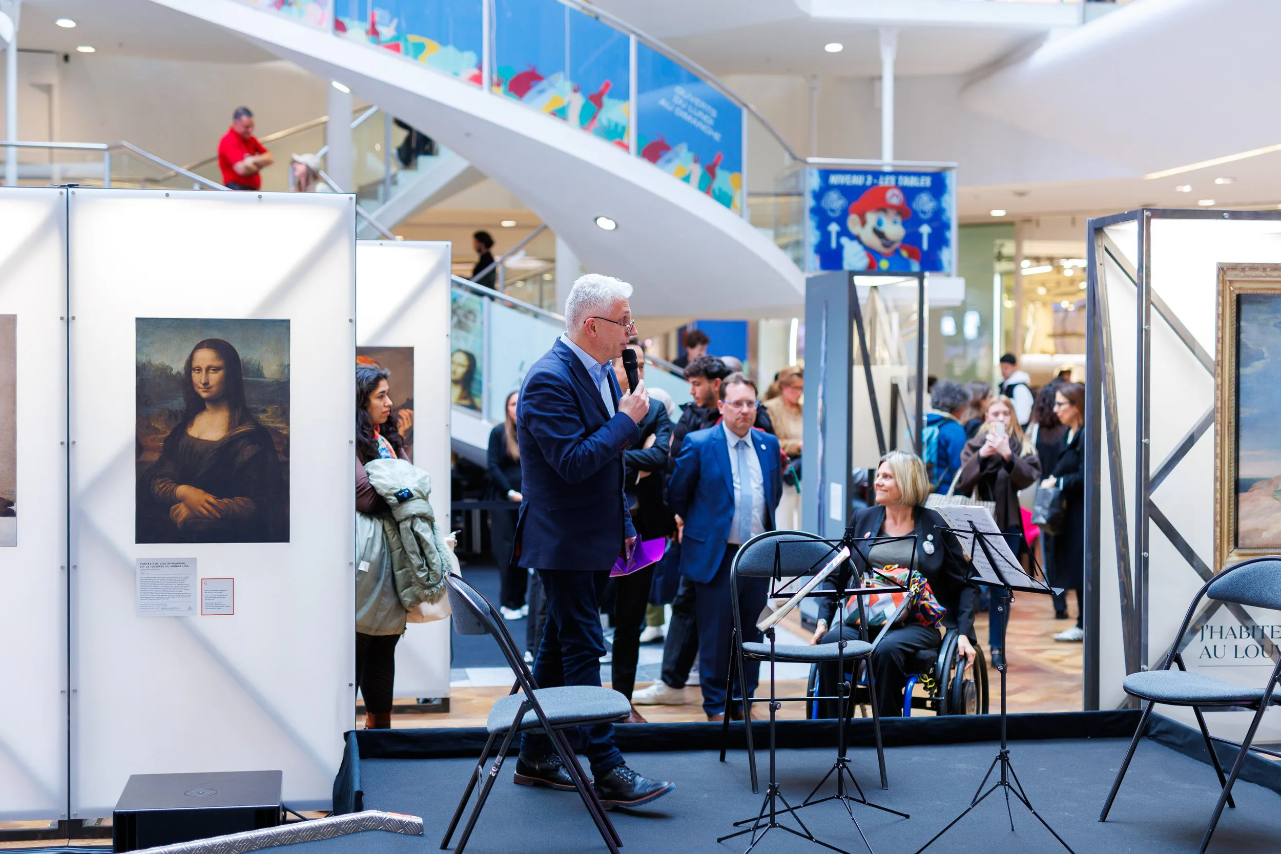 Un homme avec des cheveux gris tient un micro et parle à une foule lors d'un événement dans une galerie d'art dans un centre commercial. Autour, des personnes discutent ou regardent des œuvres d'art, notamment une reproduction de la Mona Lisa. Il y a