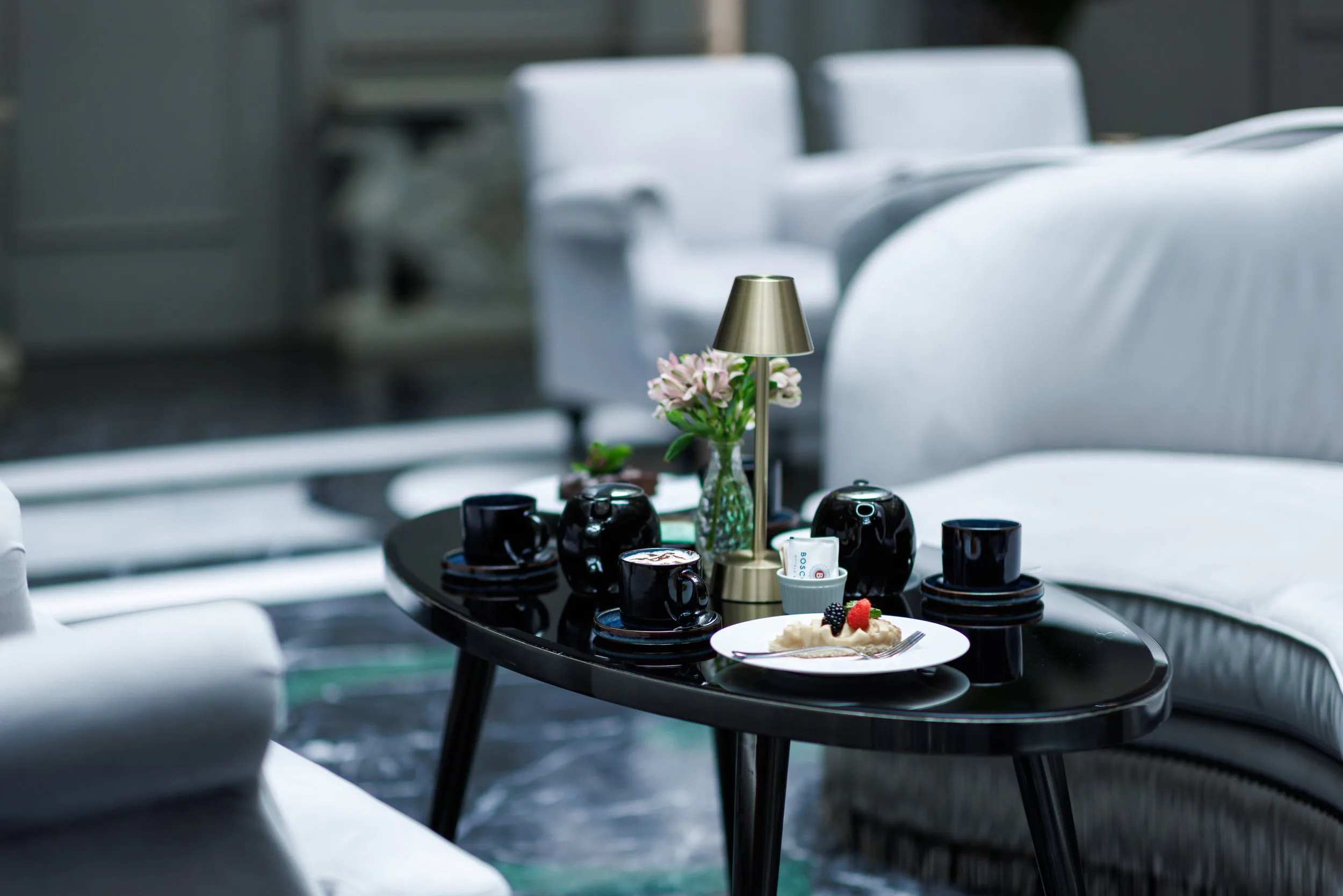 Une table noire avec une lampe dorée, une assiette avec dessert, plusieurs tasses noires, un vase avec des fleurs, dans un salon moderne avec des canapés blancs.