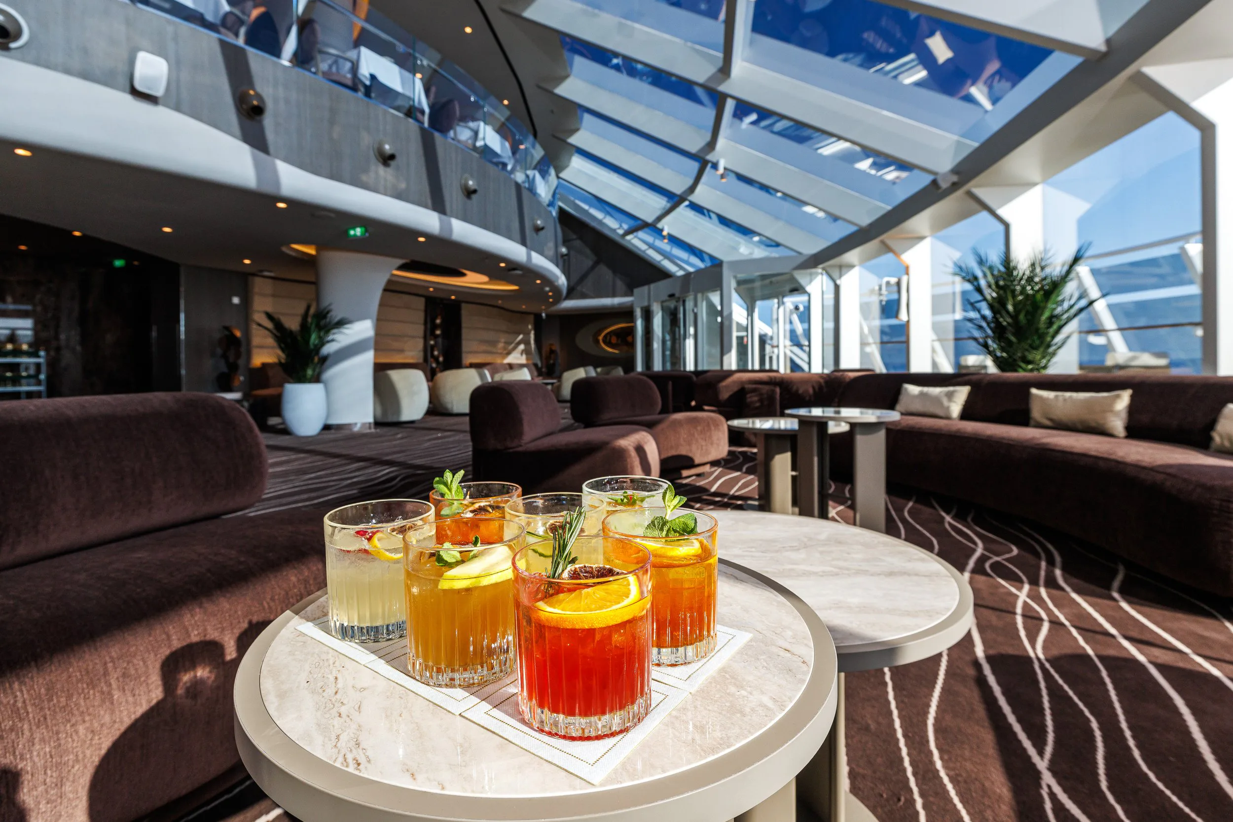 Verre de cocktails colorés sur une table dans un lounge luxueux avec de grandes fenêtres et des sièges rembourrés.