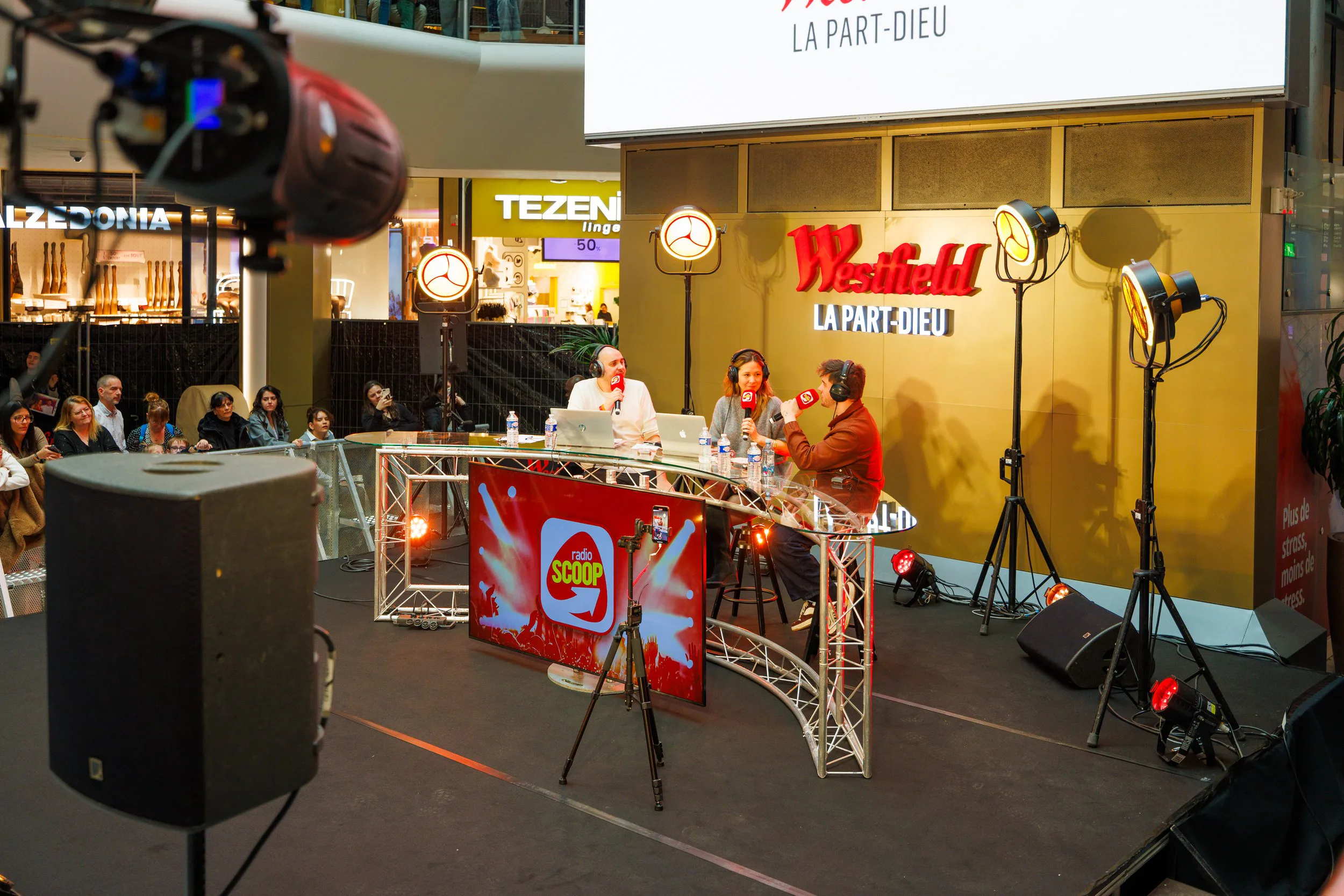 Animation en direct dans un centre commercial avec trois personnes - deux femmes et un homme, tous portant des écouteurs et tenant des microphones, discutant derrière une table aux couleurs de Radio Scoop, entourée de lumières sur un petit podium. Au