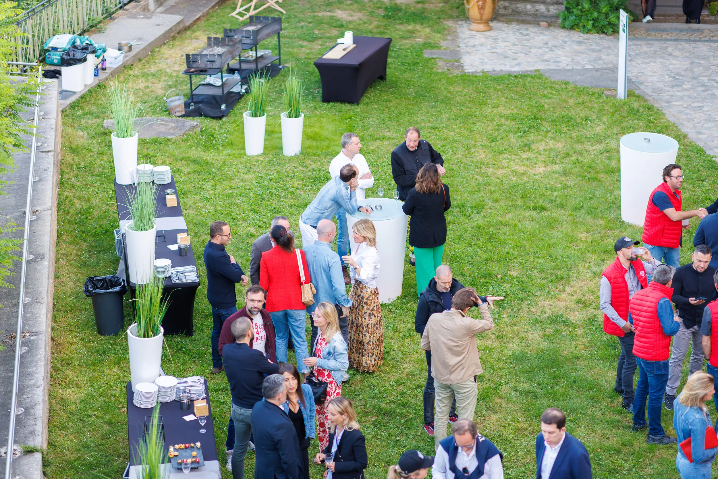 Un groupe de personnes lors d'une fête ou d'un rassemblement en plein air, avec des tables de nourriture, des boissons, et des décorations végétales. Certaines personnes discutent, d'autres prennent des photos ou se servent à boire.