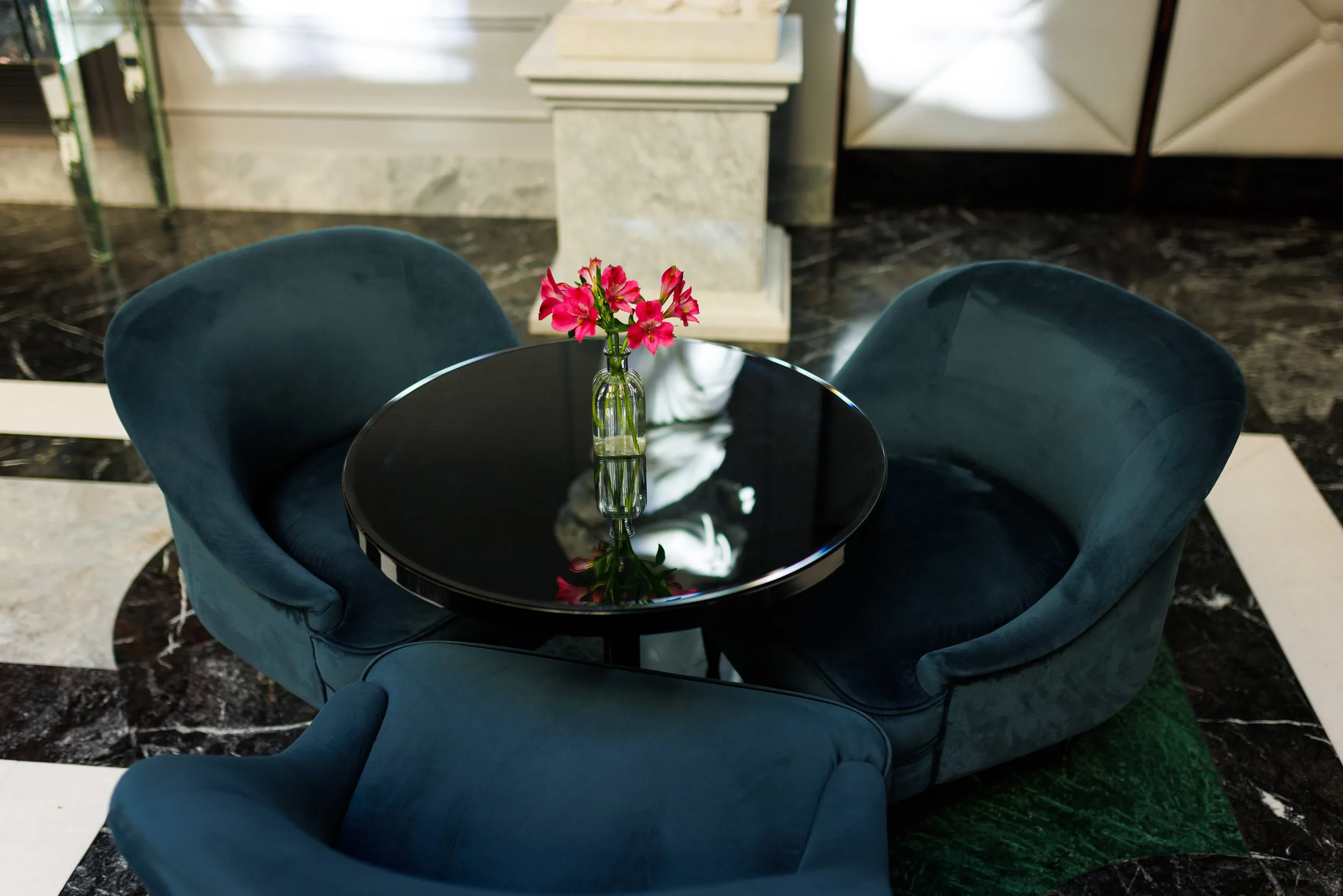 Un petit tableau rond avec une bouteille de verre contenant des fleurs roses, entouré de deux fauteuils en velours bleu foncé, dans un intérieur élégant avec un sol en marbre noir et blanc.