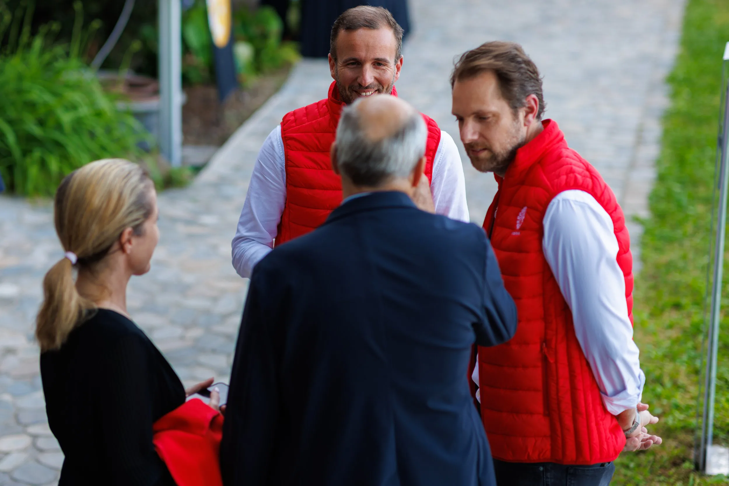 Quatre personnes discutent en cercle sur un chemin pavé extérieur, deux portent des vestes rouges, une une veste noire, une autre une chemise blanche, tous semblent engagés dans une conversation.»