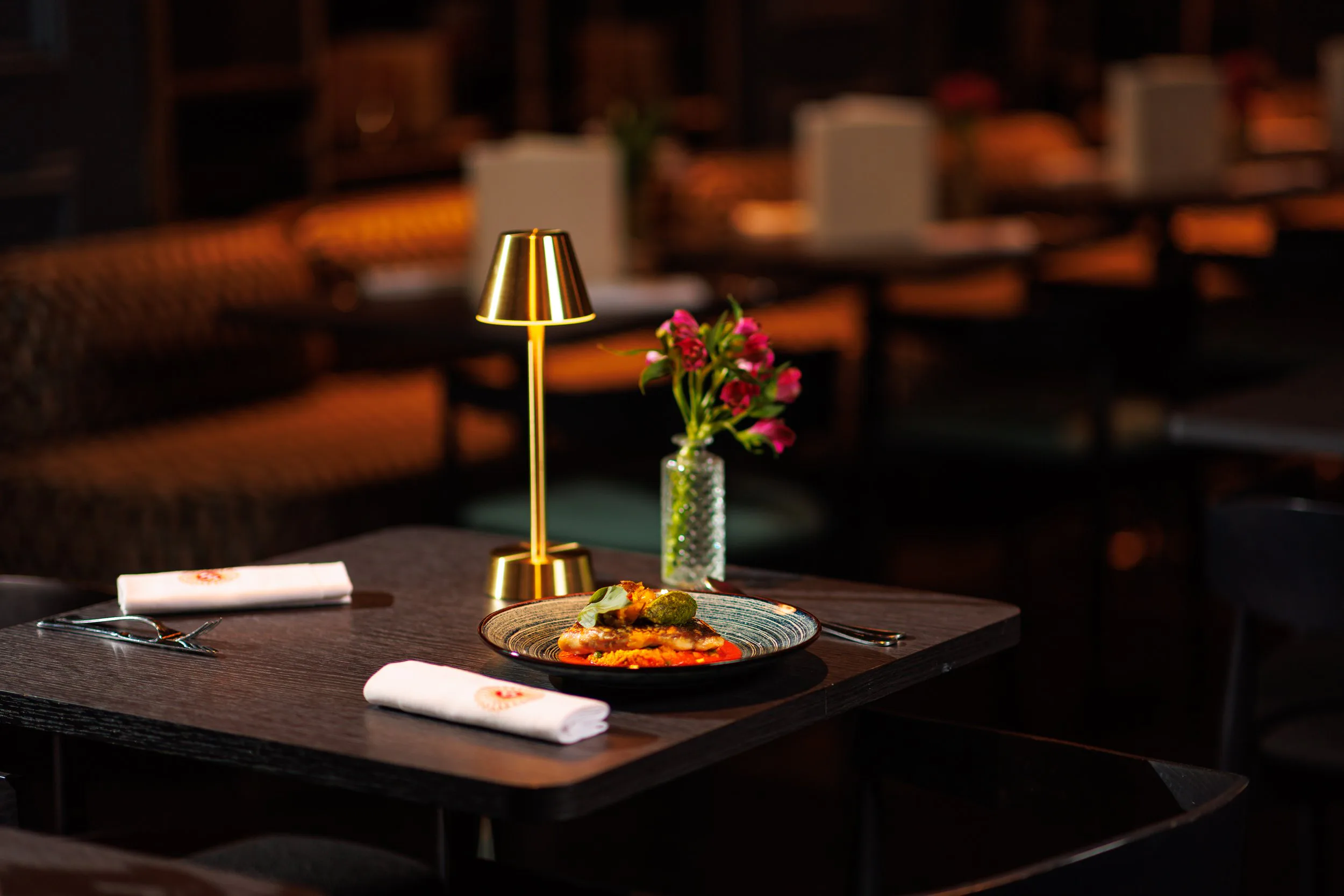 Une table de restaurant élégante avec un plat, un vase avec des fleurs roses, une lampe dorée et des couverts, ambiance chaleureuse.