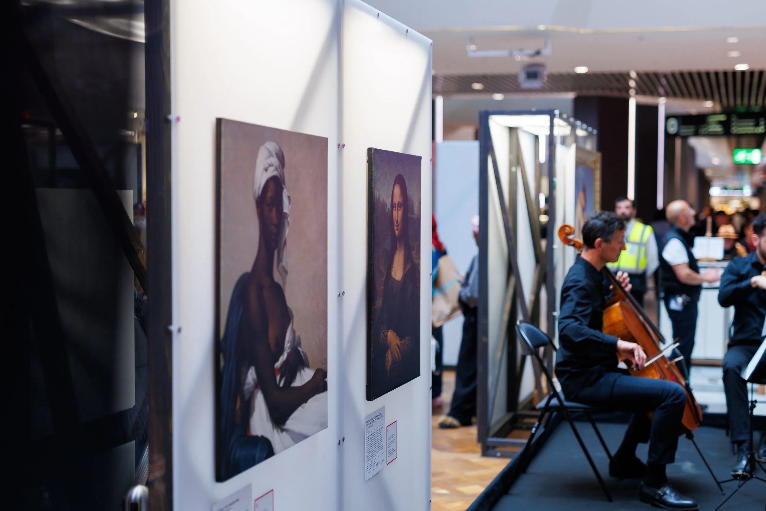 Une exposition d'art avec des tableaux accrochés à un mur blanc, et un groupe de musiciens jouant du violon en arrière-plan dans un espace intérieur.