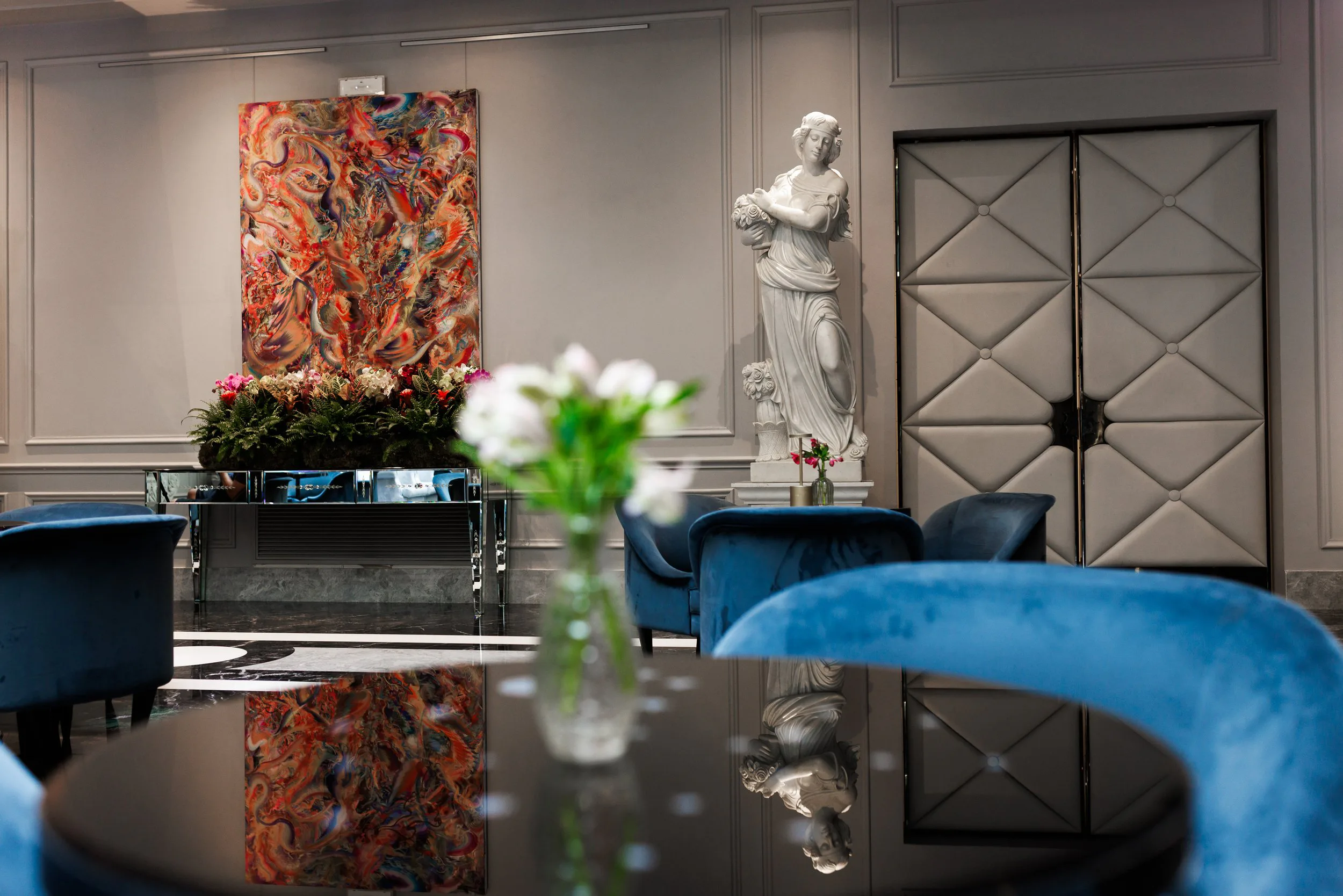 Intérieur d'un salon élégant avec une sculpture en marbre d'une femme, un tableau abstrait coloré, des fauteuils en velours bleu, des fleurs dans un vase et des murs gris avec des panneaux panelés.