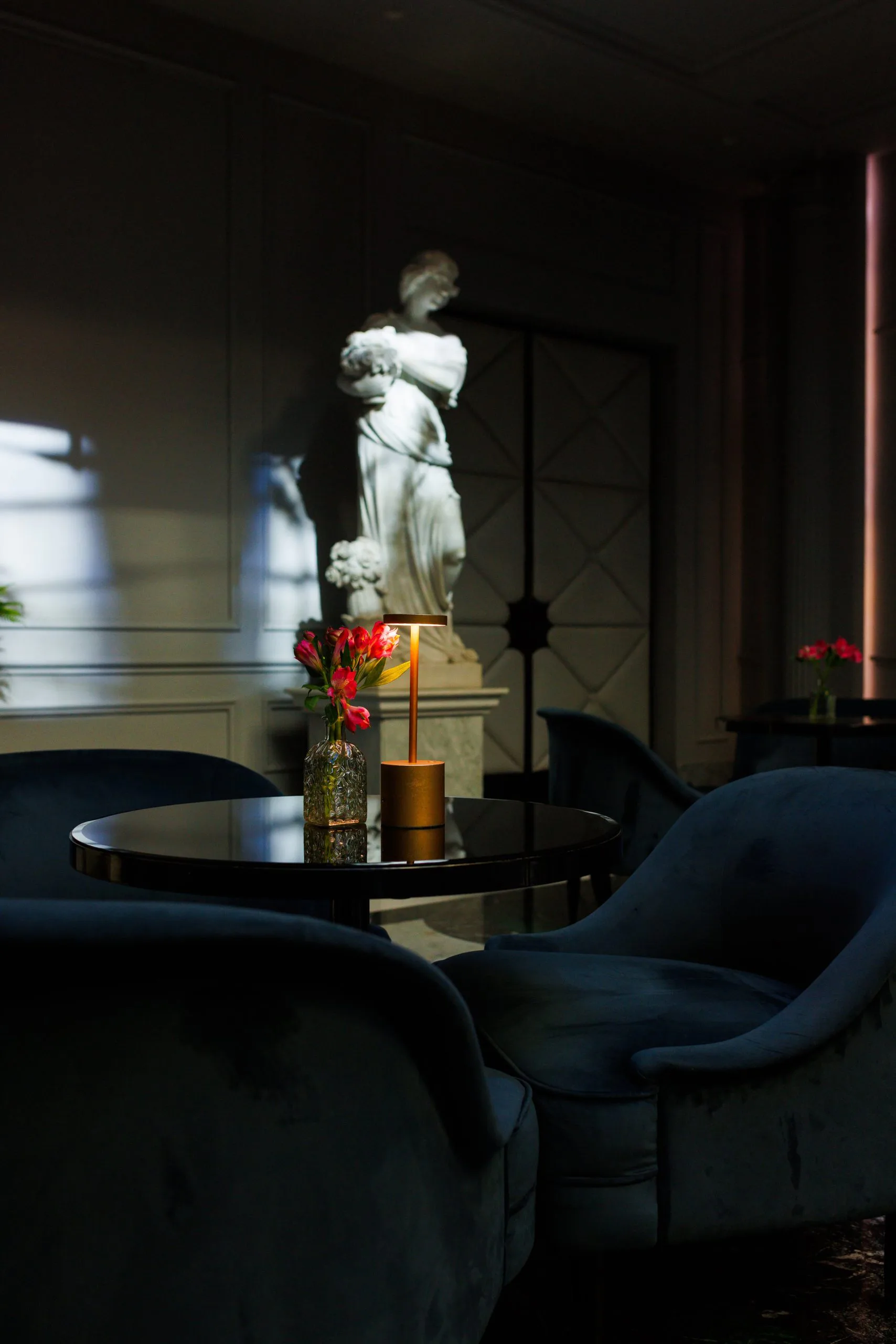 Une pièce élégante avec un tableau noir, un vase avec des fleurs roses, une lampe, un fauteuil et une grande statue de marbre représentant une femme antique.