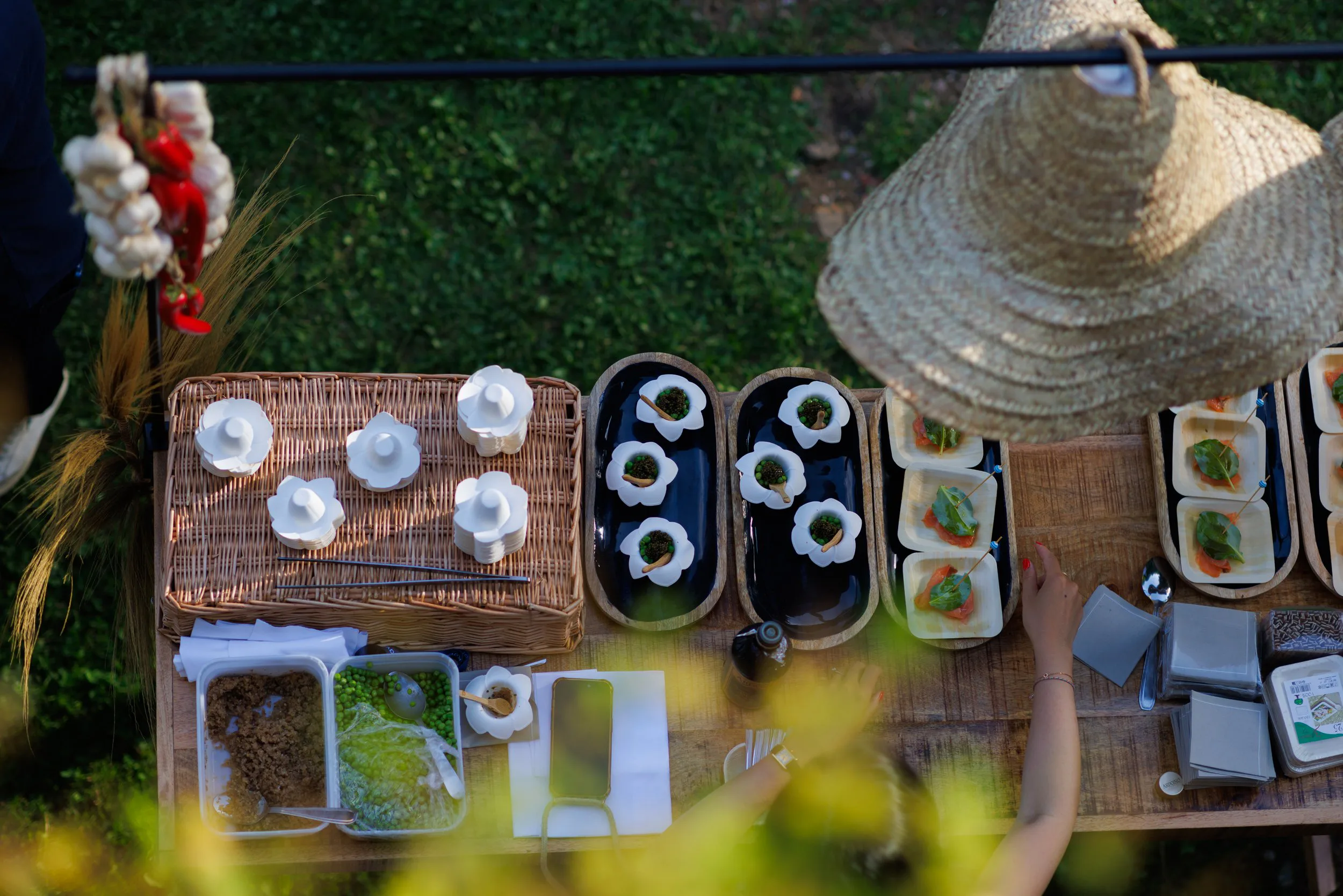 Table de buffet en plein air avec des petites assiettes de nourriture, des couverts, un chapeau de paille et des décorations en feuilles, vue en hauteur.