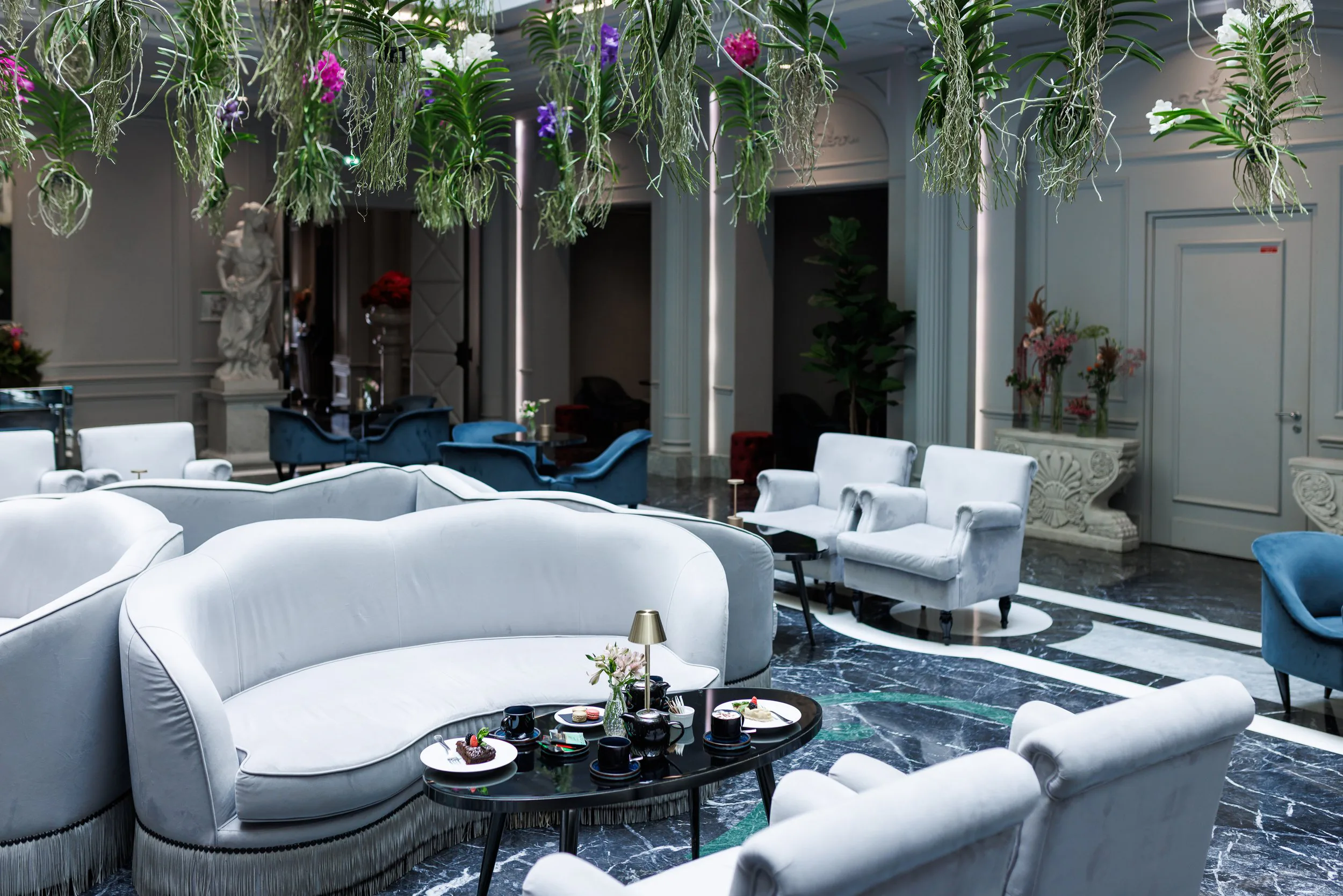 Salon élégant avec canapés en velours blanc, tables basses avec boissons et desserts, décor de plantes suspendues et statues en arrière-plan.