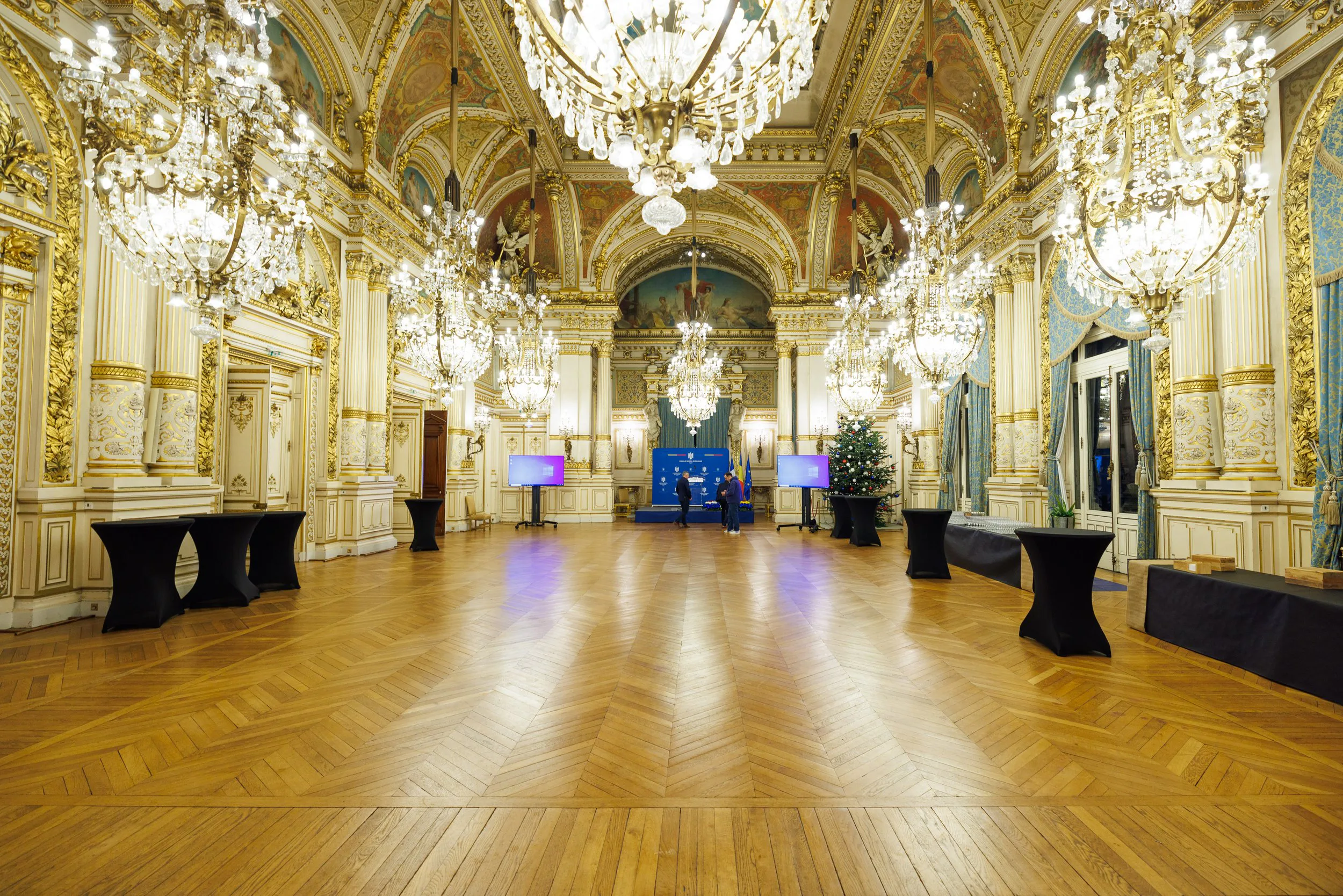 Une salle de réception somptueuse avec un plafond orné de fresques et plusieurs lustres en cristal. La pièce a des murs décorés de moulures dorées, des fenêtres avec des rideaux bleus, et un sapin de Noël décoré. Il y a des tables noires dispersées d