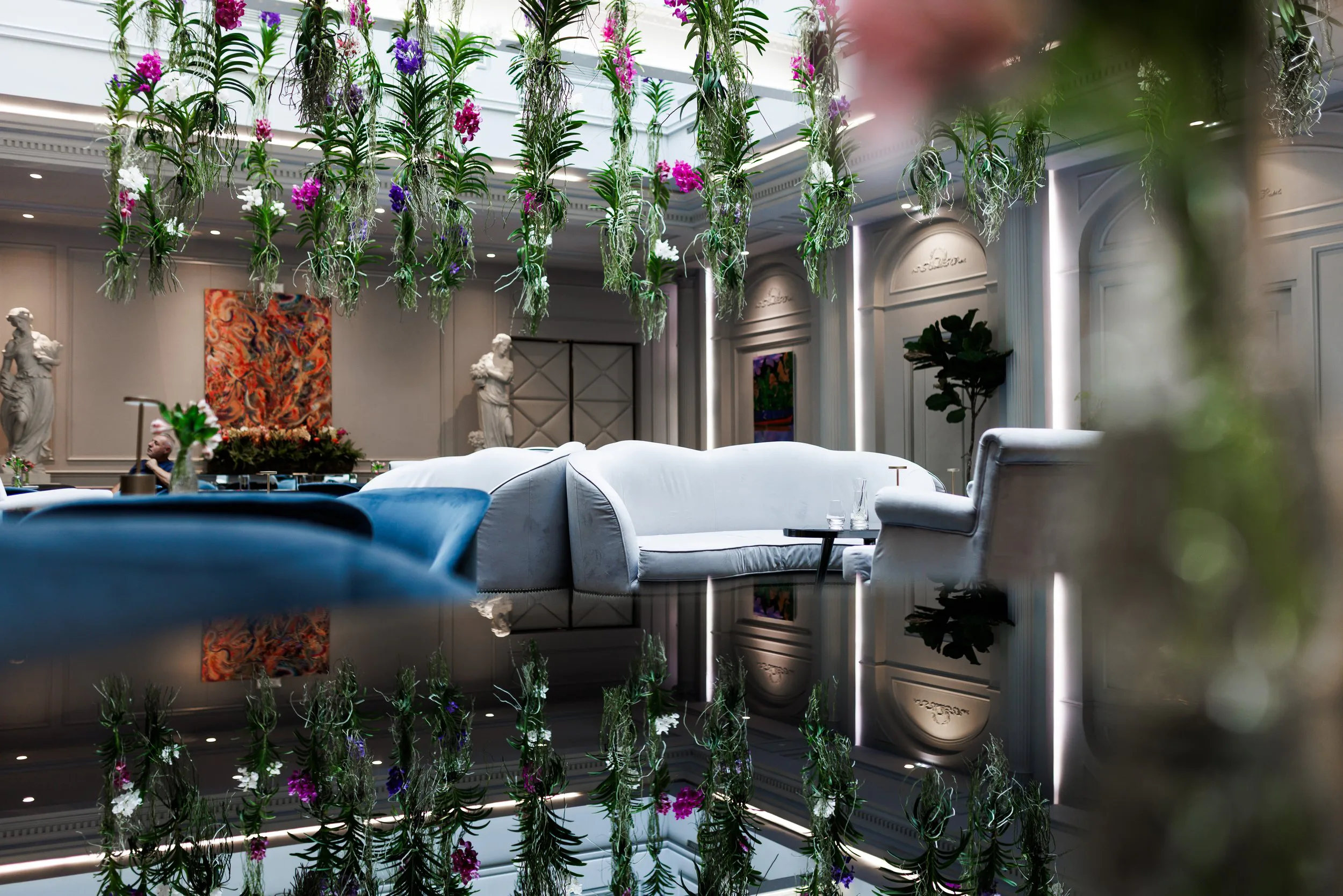 Intérieur d'un espace lounge élégant avec des canapés blancs et bleus, des sculptures classiques, des œuvres d'art colorées sur les murs, et une décoration florale suspendue en hauteur.