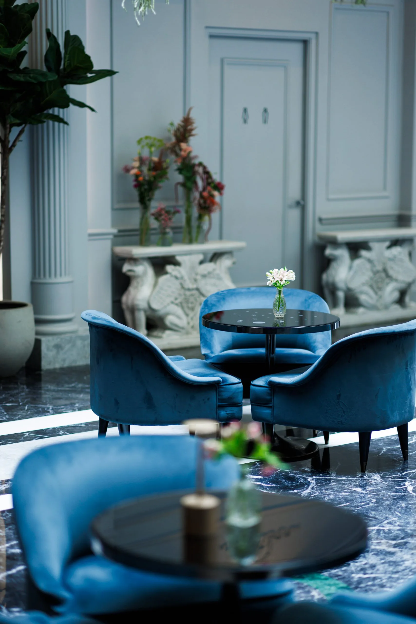 Salon élégant avec fauteuils en velours bleu, petit vase avec fleurs sur une table noire, décor intérieur avec colonnes et sculptures.