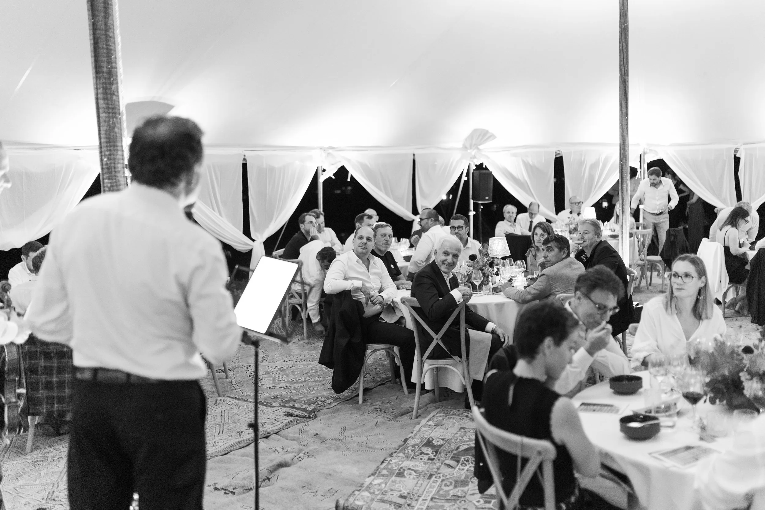 Un dîner en plein air avec plusieurs personnes assises autour de tables, un homme en avant-plan derrière un pupitre semble faire un discours, ambiance élégante et festive.