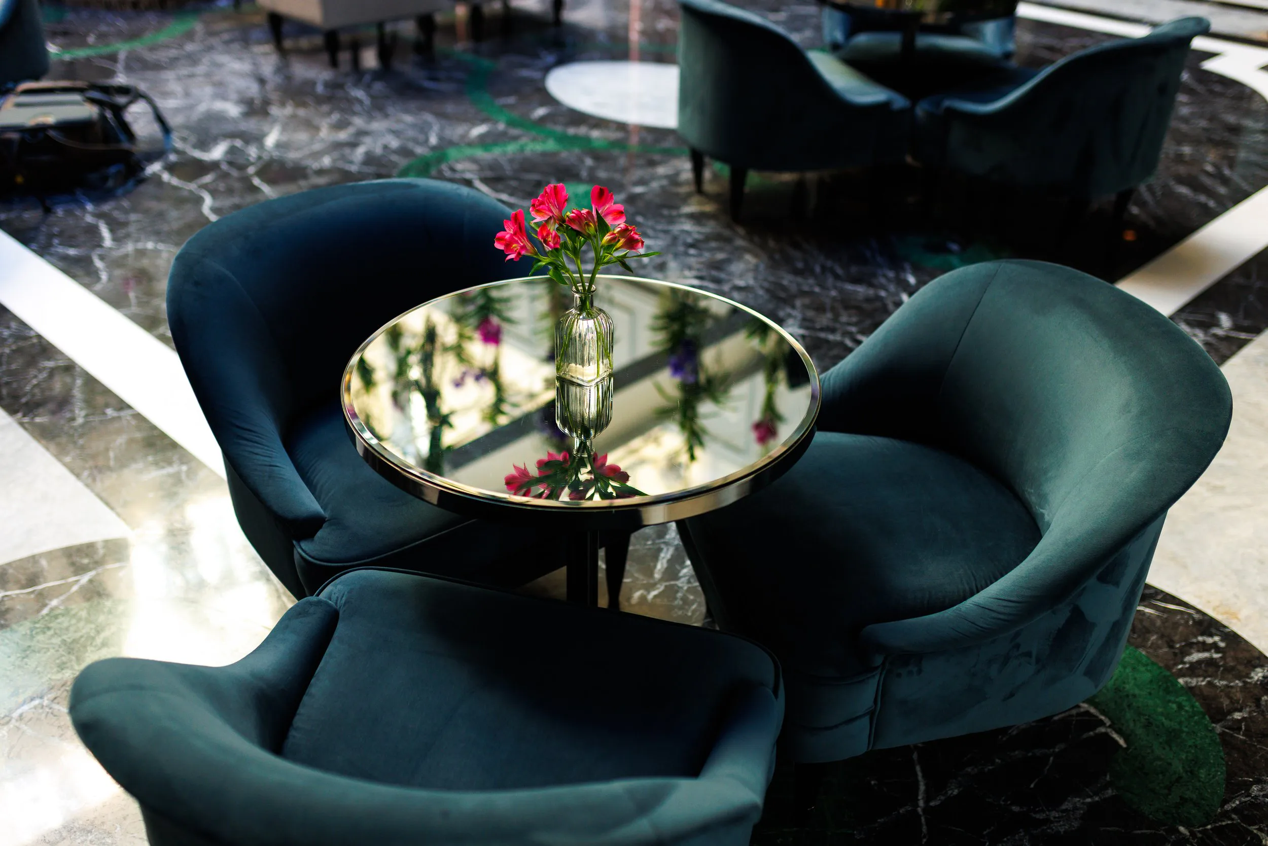 Table ronde en verre avec vase de fleurs roses, entourée de trois fauteuils verts en velours, dans un espace intérieur élégant.