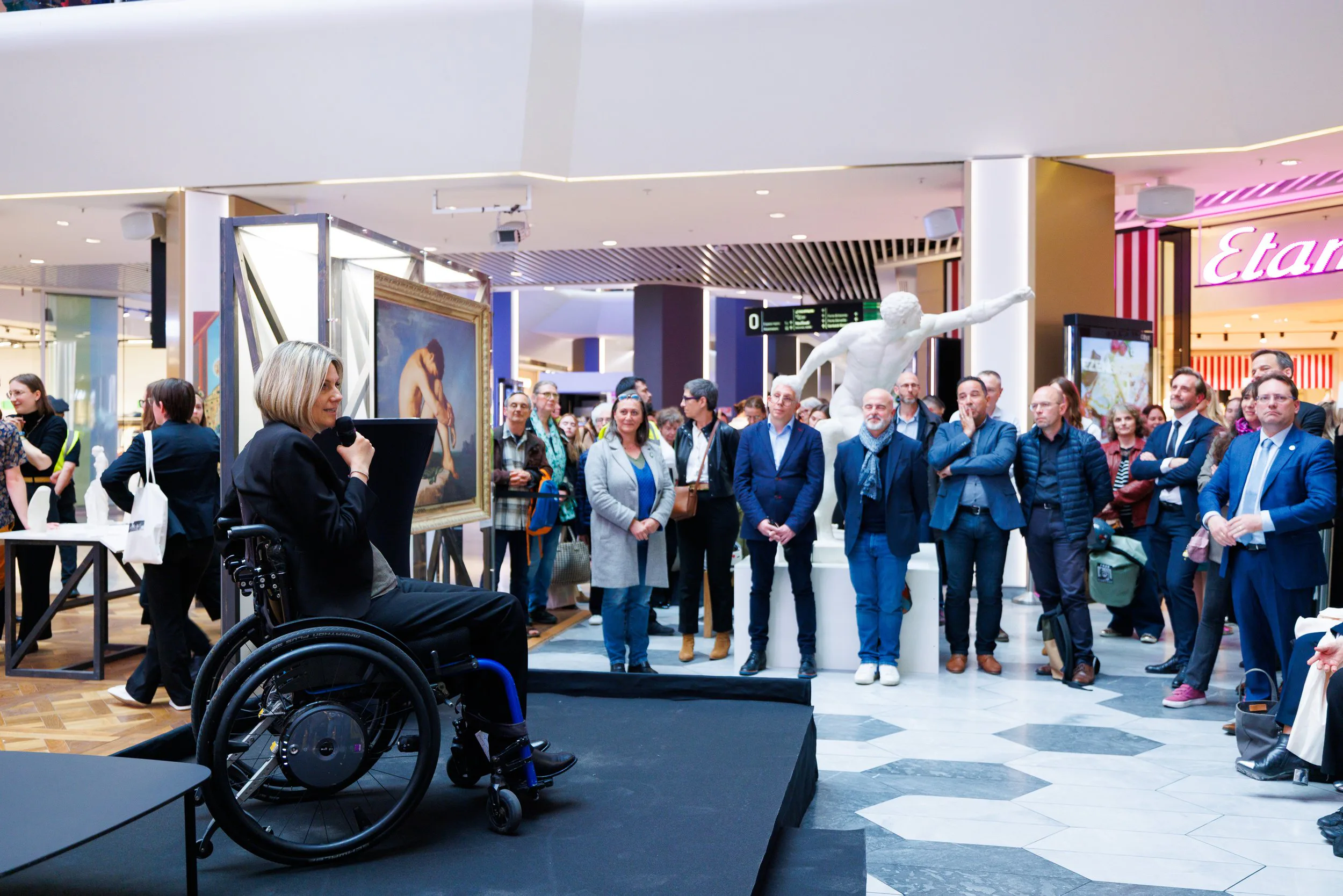 Une femme en fauteuil roulant parle dans un micro lors d'une présentation ou d'une cérémonie dans un centre commercial, avec une audience de personnes regardant et écoutant attentivement.