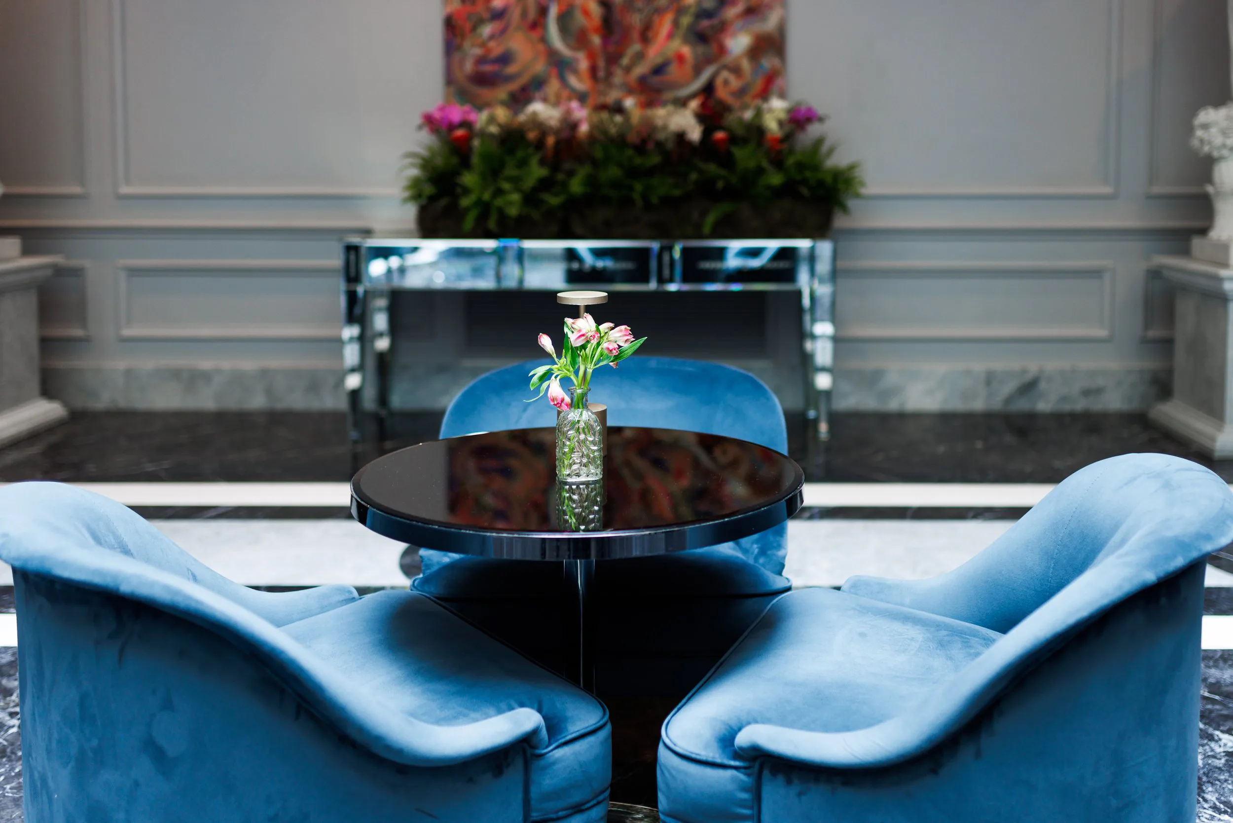 Une pièce élégante avec un meuble en miroir, un tableau abstrait, et deux fauteuils en velours bleu autour d'une table ronde en verre avec un vase de fleurs roses.