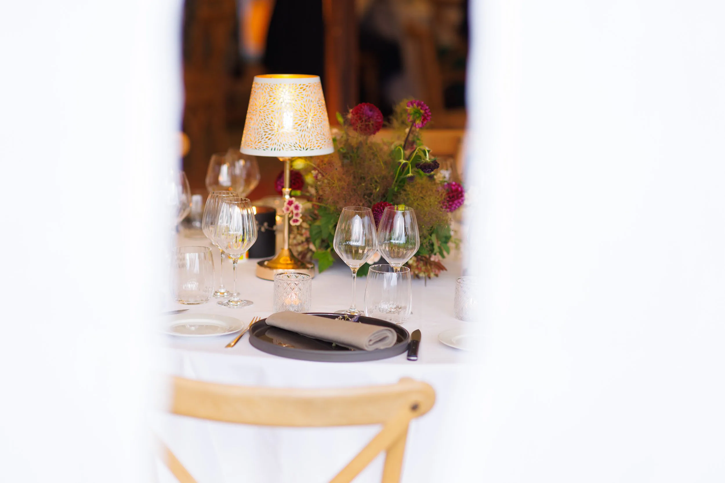 Table de dîner élégante avec vaisselle, verres à vin, bougie et bouquet de fleurs, vu à travers un espace entre deux murs blancs.