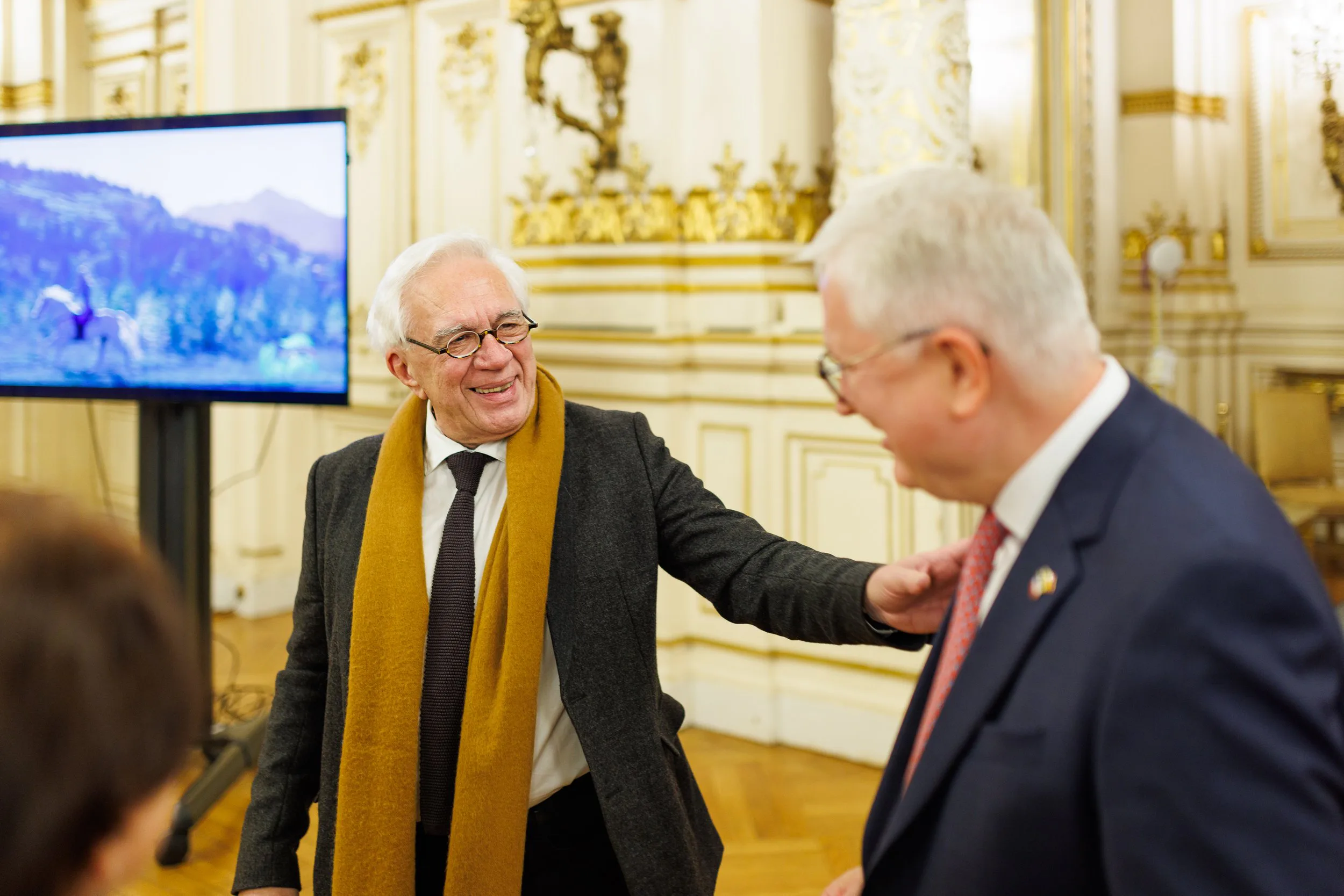 Deux hommes d'affaires souriants conversent dans une salle ornée de dorures, avec un grand écran à l'arrière affichant une image de montagne et un cheval