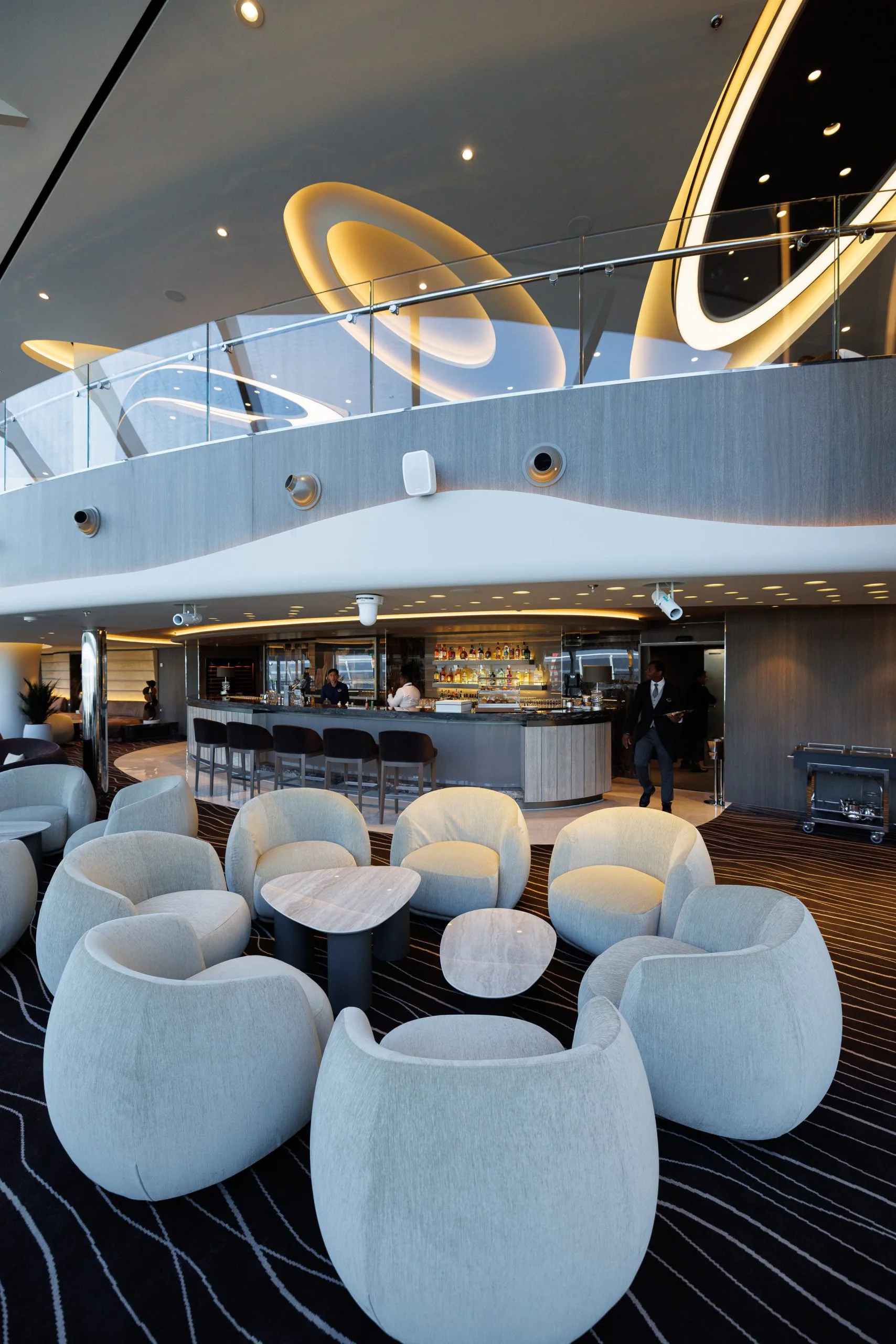 Intérieur d'un lounge avec des fauteuils en tissu beige et une table en marbre. En arrière-plan, un bar avec des barmen, et un espace en mezzanine avec un design moderne et des éclairages artistiques au plafond.