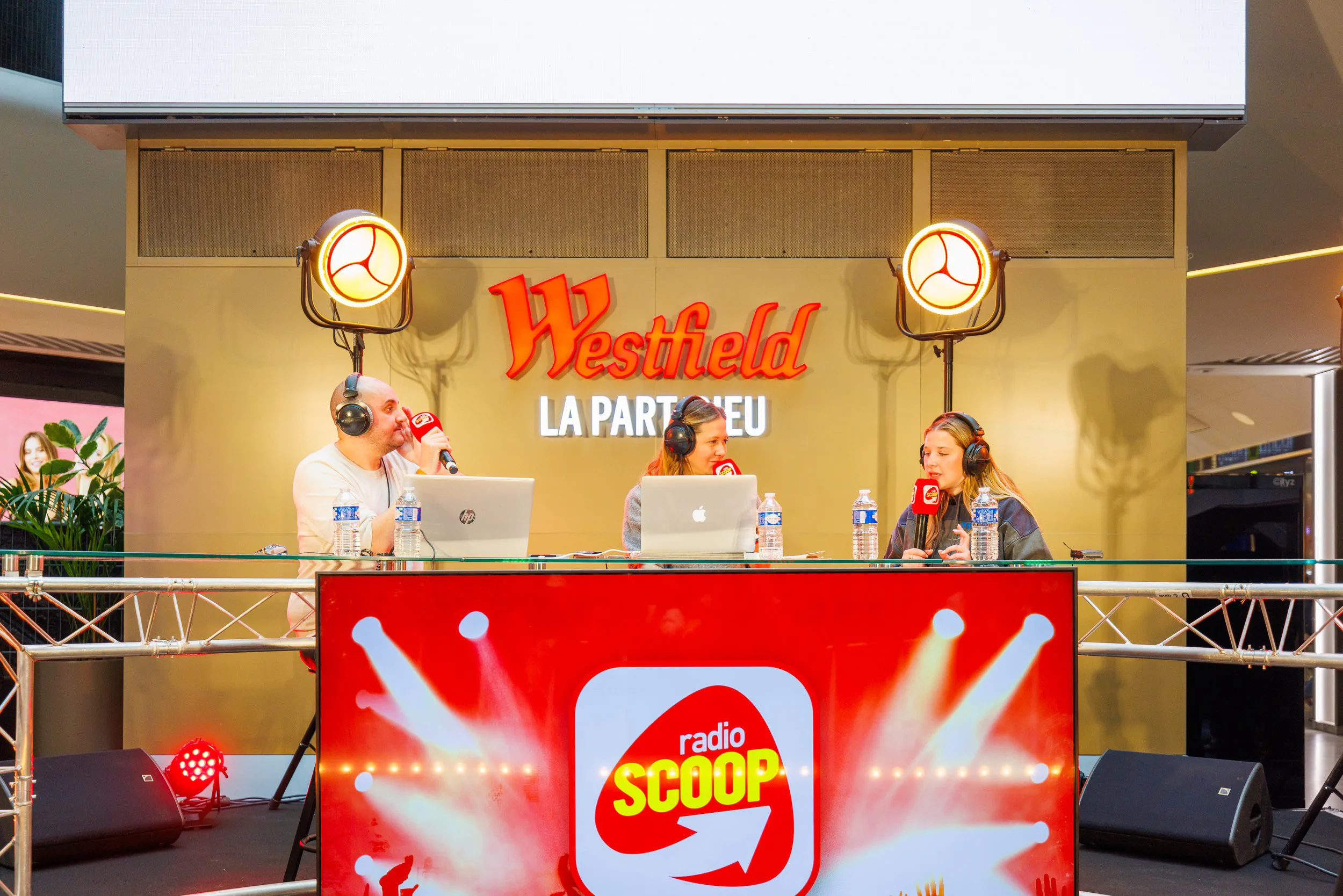 Trois personnes écoutant une émission en direct à la radio Radio Scoop, avec microphones et casques, dans un exposé Public à l'intérieur d'un centre commercial. Le fond affiche un panneau Westfield La Part Dieu avec deux lumières suspendues.