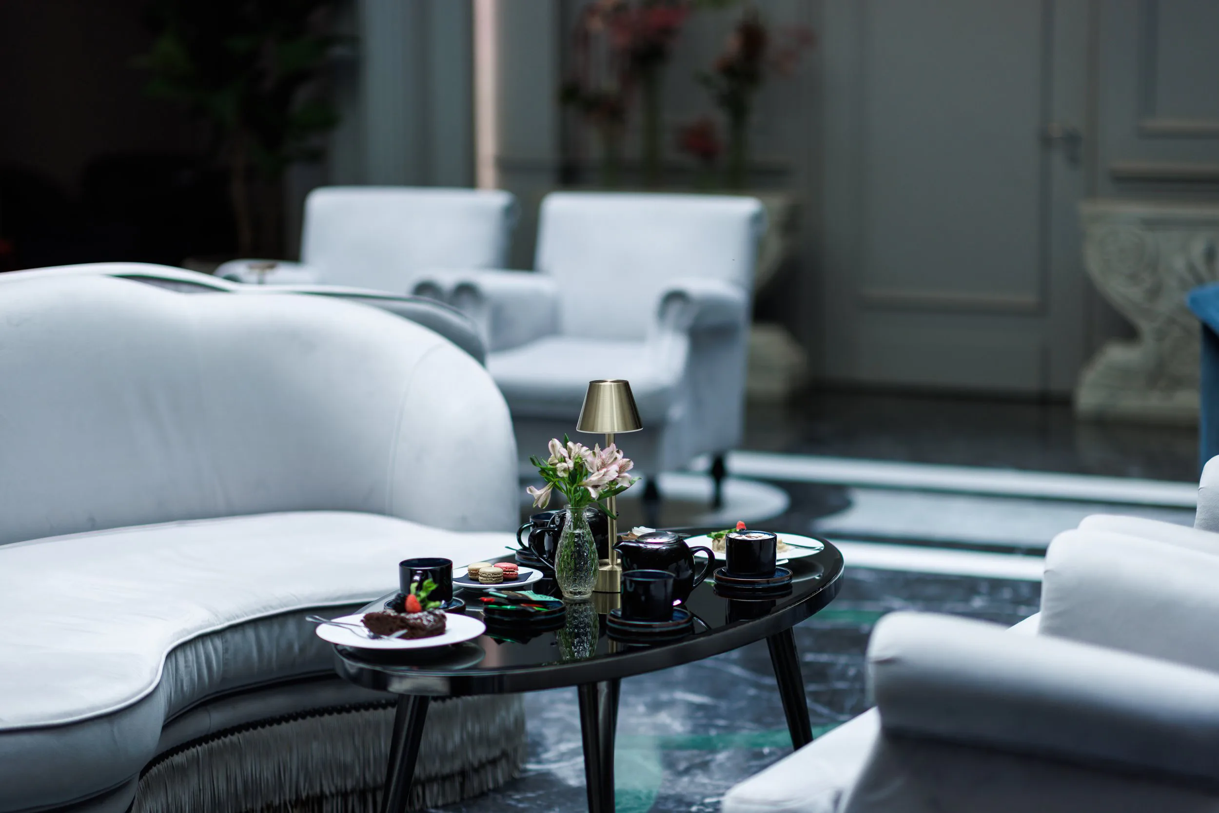 Salon élégant avec mobilier blanc, table noire avec vaisselle et décor floral, lampe dorée.