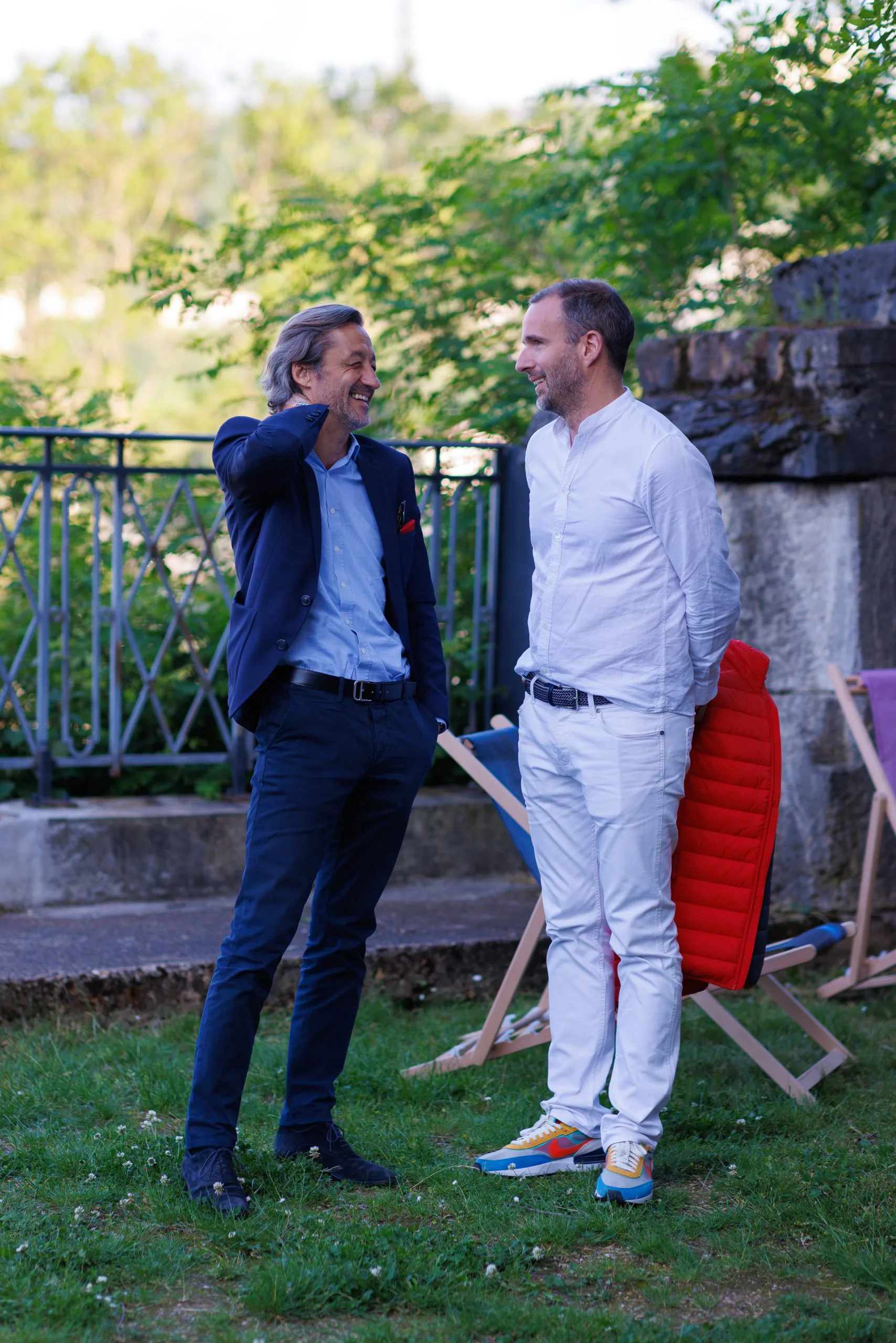Deux hommes souriants discutent dans un jardin, l'un en costume foncé, l'autre en vêtements décontractés blancs, avec des chaises longues en arrière-plan.