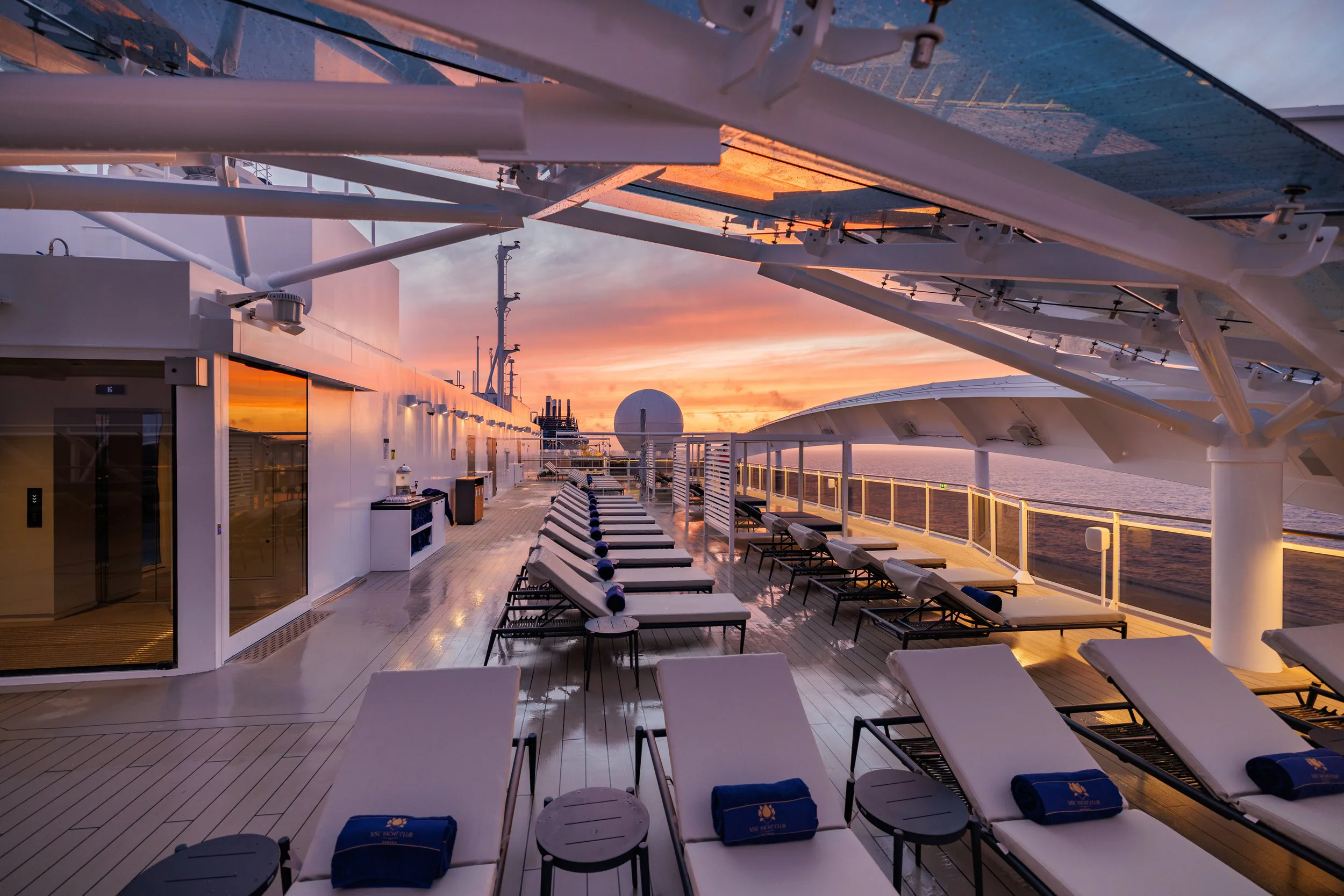 Vue d'un pont de croisière au coucher de soleil, avec des chaises longues blanches, une échelle, et une mer calme à l'arrière-plan.