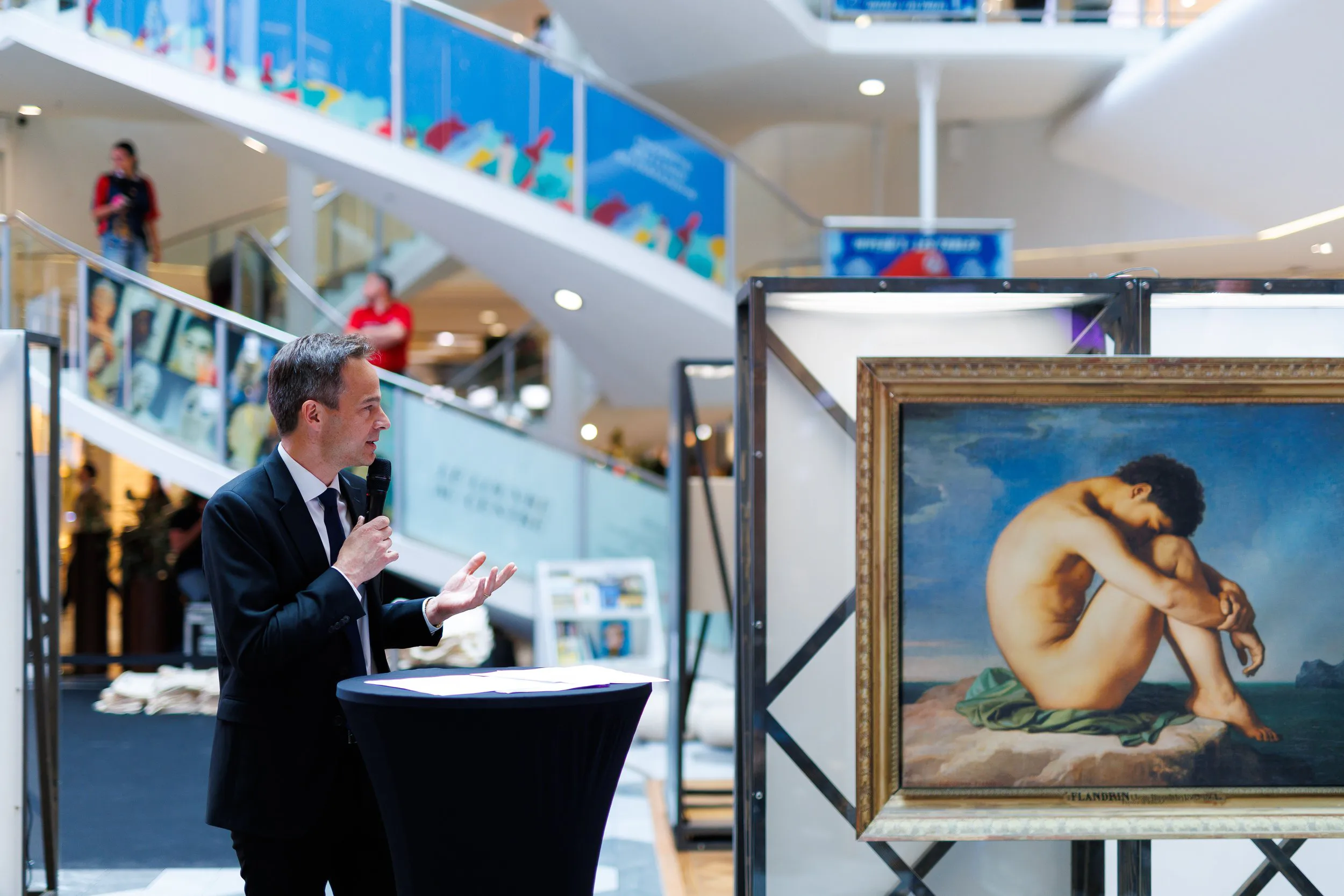 Homme en costume noir parlant avec un micro dans un centre commercial, près d'une peinture représentant un homme nu assis, avec une toile de fond de ciel et de paysage marine.